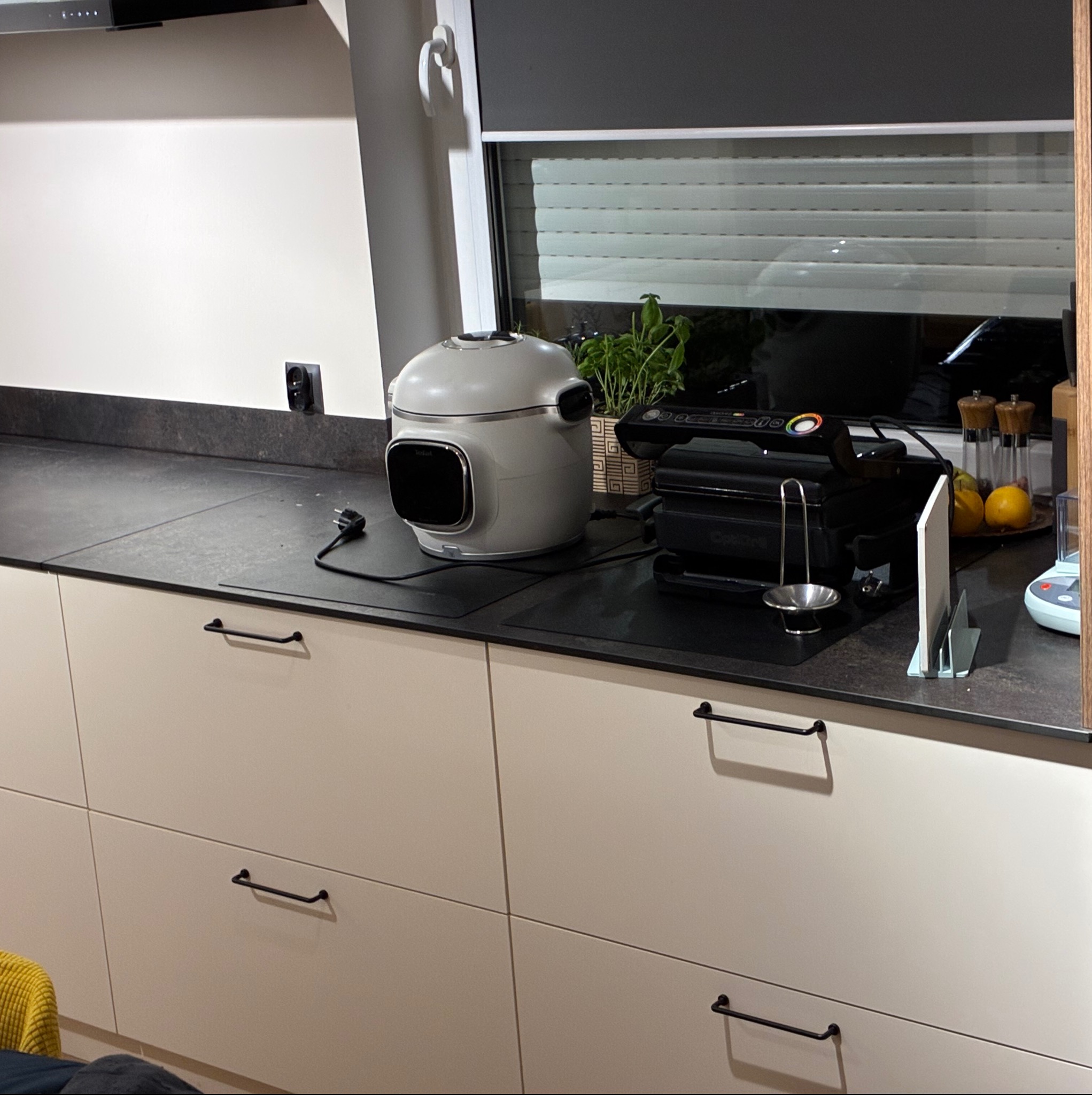 Jasna kuchnia z beżowymi frontami szafek i czarnymi uchwytami. Na blacie stoją urządzenia kuchenne i przyprawy. W tle okno z roletą.