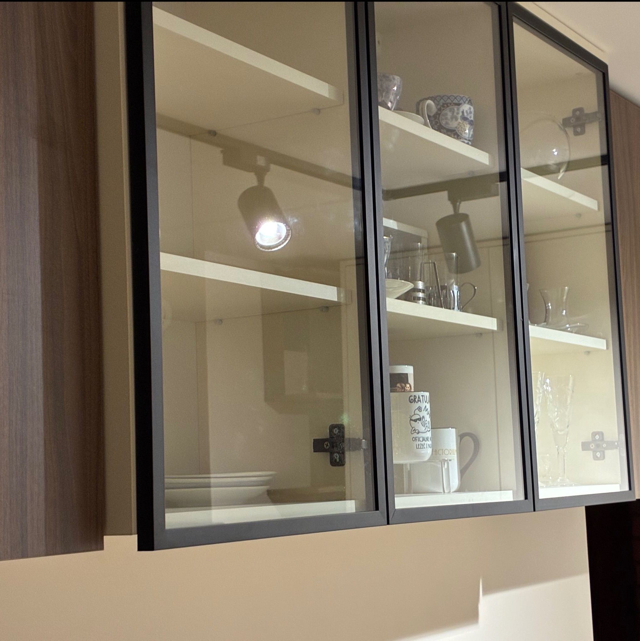 Szklana szafka wisząca z czarnymi ramami, oświetleniem punktowym i naczyniami w środku. Widoczne detale mocowań i półek. Ujęcie z bliska, prezentujące estetykę i funkcjonalność.