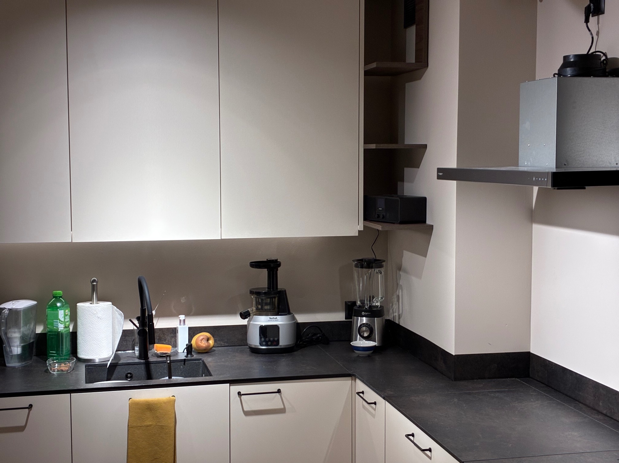 Nowoczesna kuchnia z białymi szafkami, czarnym blatem i sprzętami AGD: wyciskarka, blender. Minimalistyczny design z otwartymi półkami i okapem.