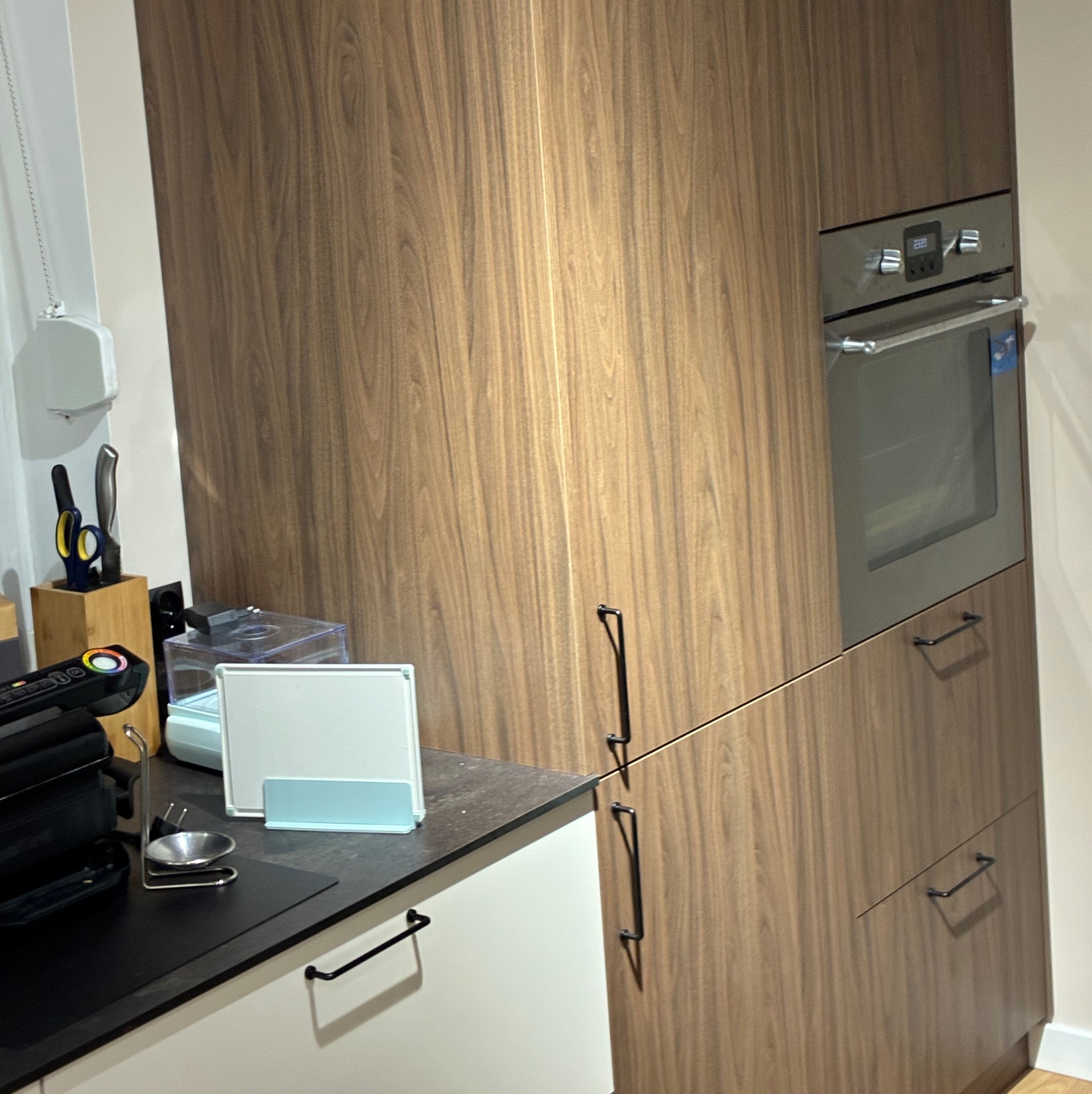 Nowoczesna zabudowa kuchenna z drewnianymi frontami i wbudowanym piekarnikiem. Na blacie akcesoria kuchenne. Minimalistyczny design z czarnymi uchwytami.