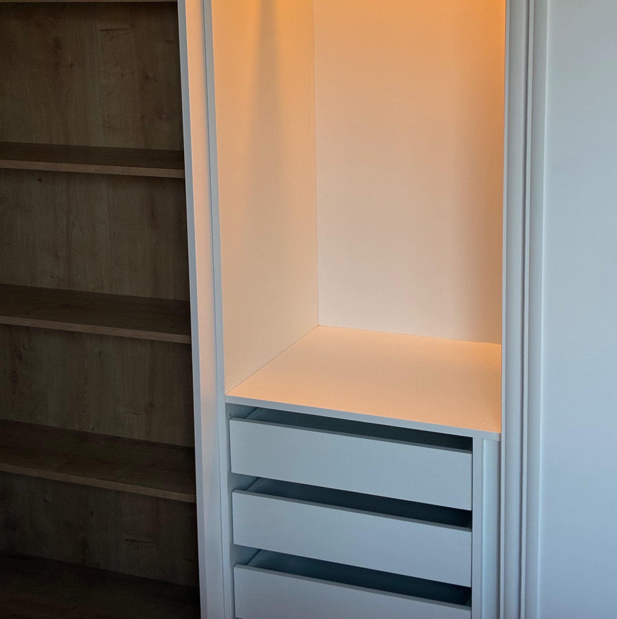 Wnętrze szafy wnękowej z szufladami i półkami, oświetlone ciepłym światłem. Widoczna struktura drewna na półkach po lewej stronie.