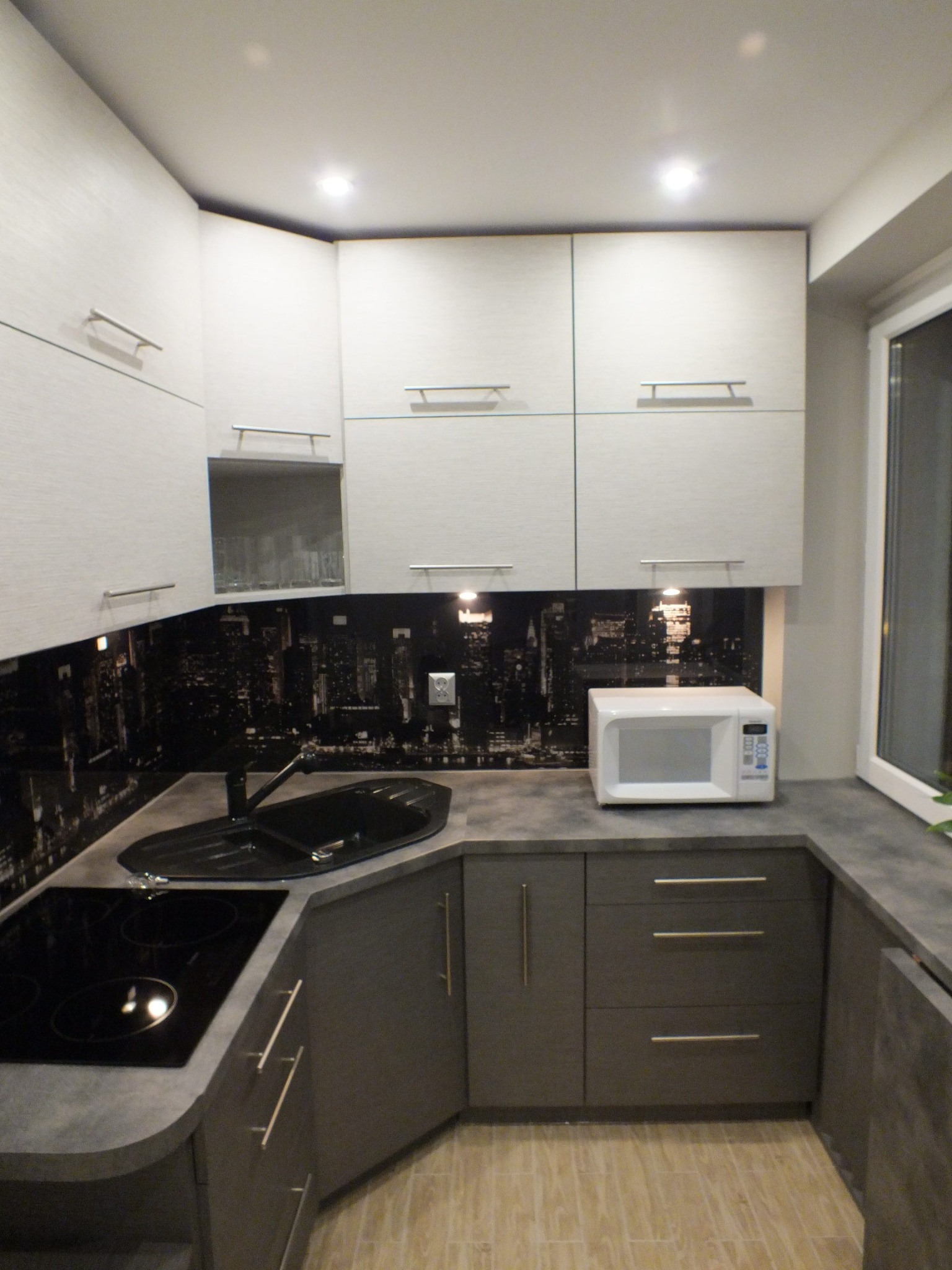 Nowoczesna kuchnia w odcieniach szarości i bieli z blatem imitującym beton. Nad blatem panel z motywem panoramy miasta. Widoczna płyta indukcyjna i mikrofalówka.