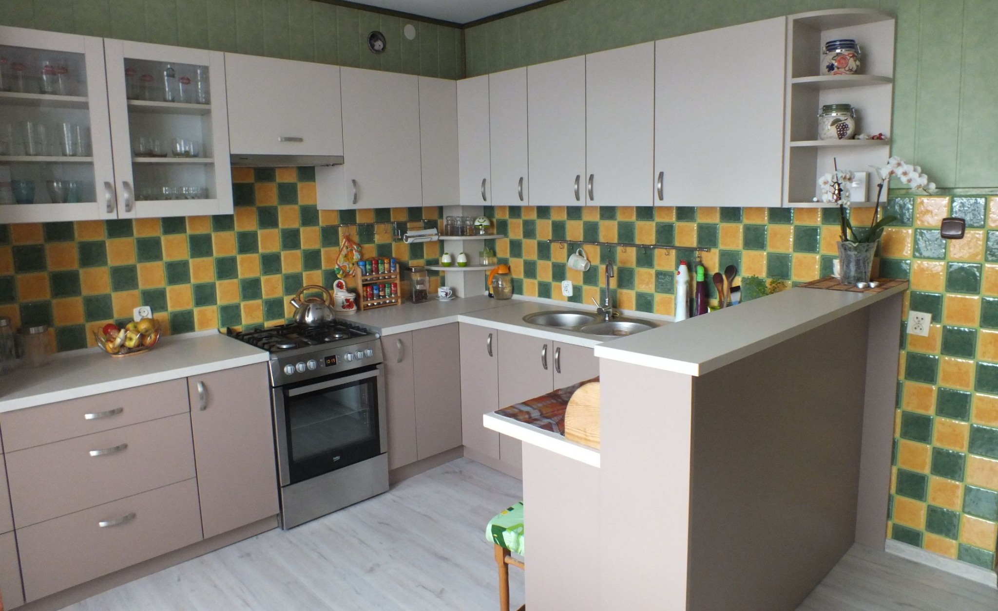 Jasna kuchnia w kształcie litery L z białymi szafkami górnymi i beżowymi dolnymi, z blatem w kolorze złamanej bieli i mozaiką z zielono-żółtych płytek na ścianie.