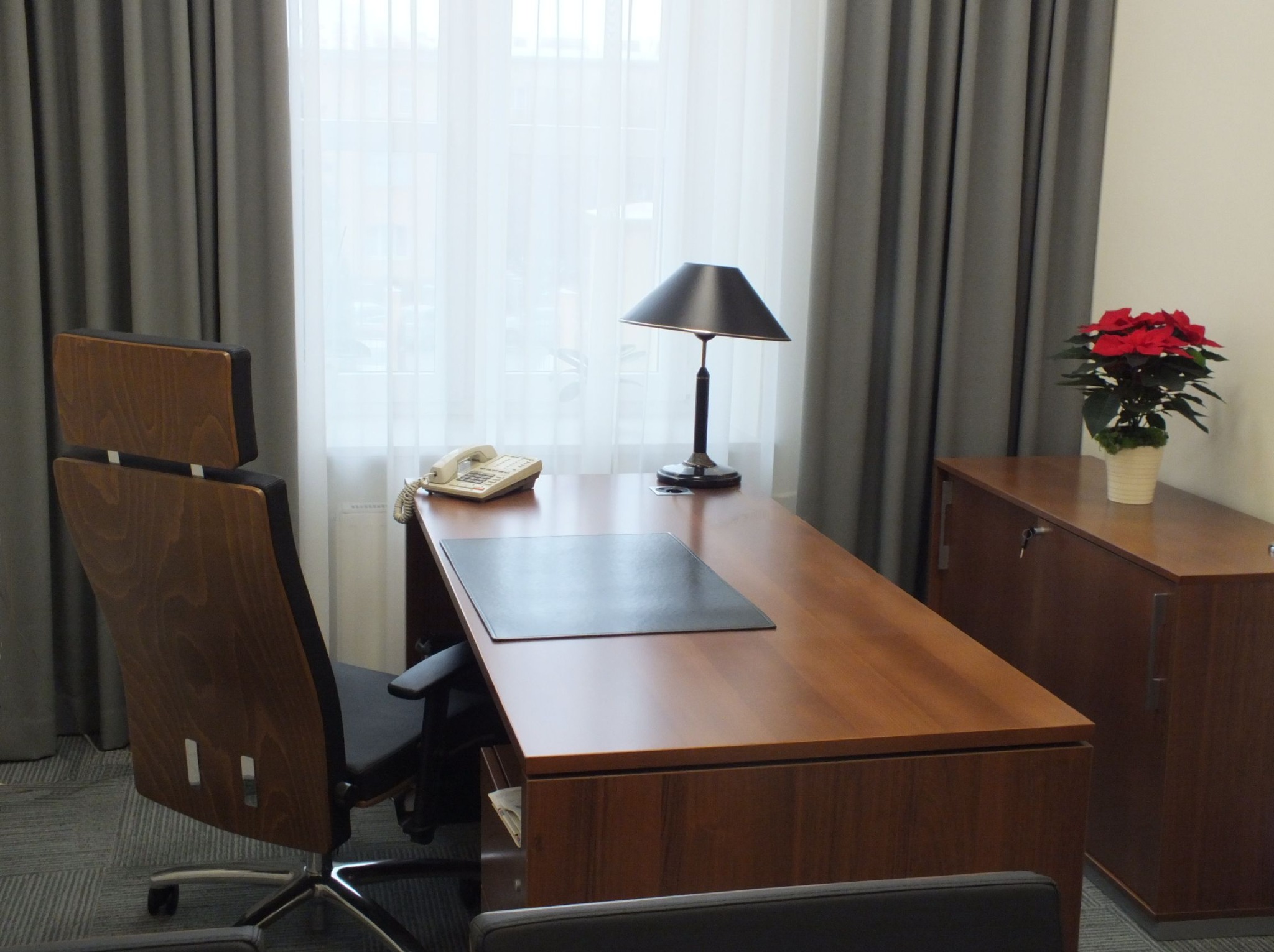 Biuro z drewnianym biurkiem, krzesłem obrotowym, lampką i szafką. Na biurku telefon i podkładka. W tle zasłony i kwiat doniczkowy. Stonowana kolorystyka.