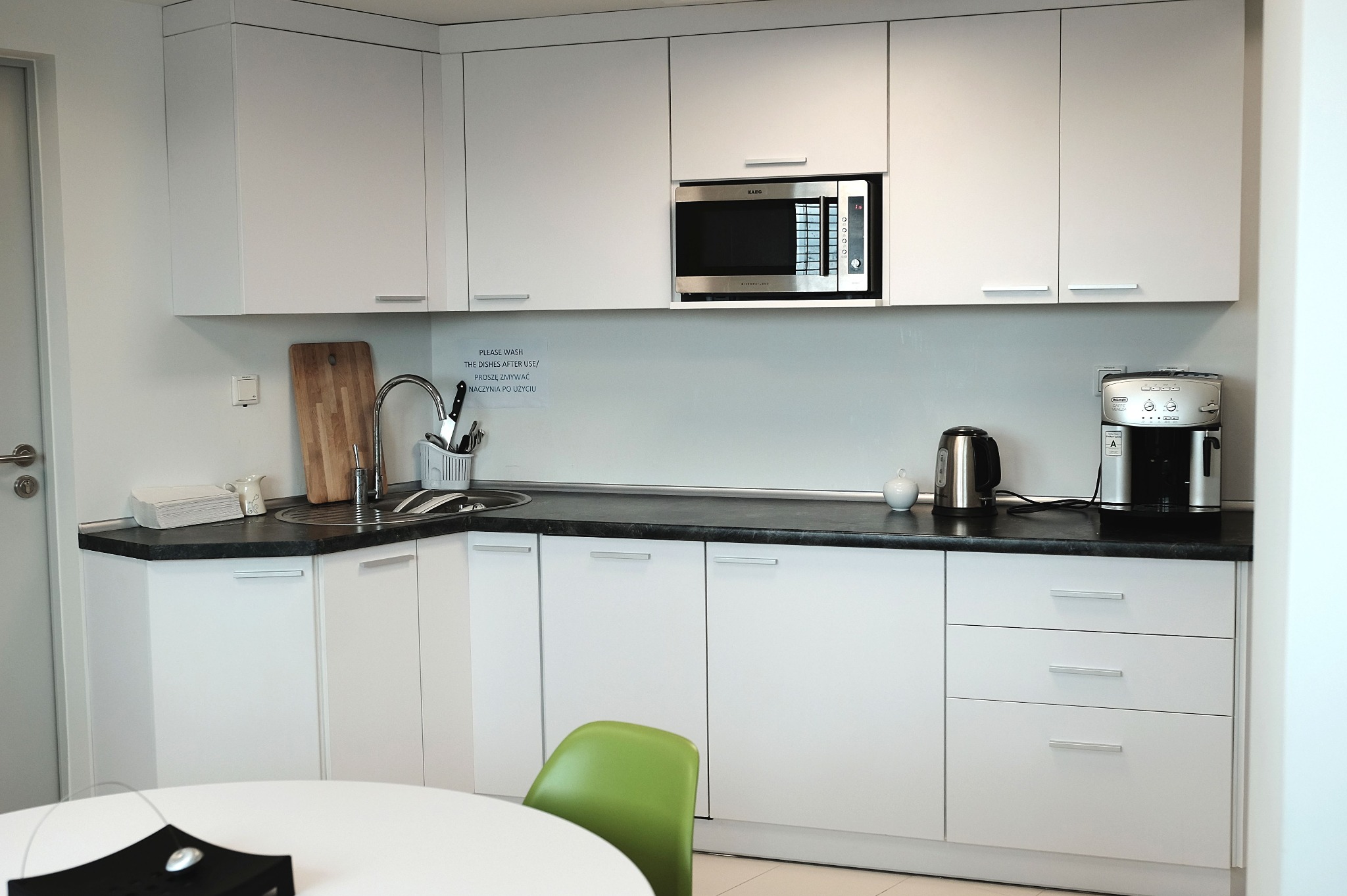 Jasna kuchnia z białymi szafkami i czarnym blatem. Na blacie zlewozmywak, mikrofalówka i ekspres do kawy. Widoczny fragment stołu z zielonym krzesłem.