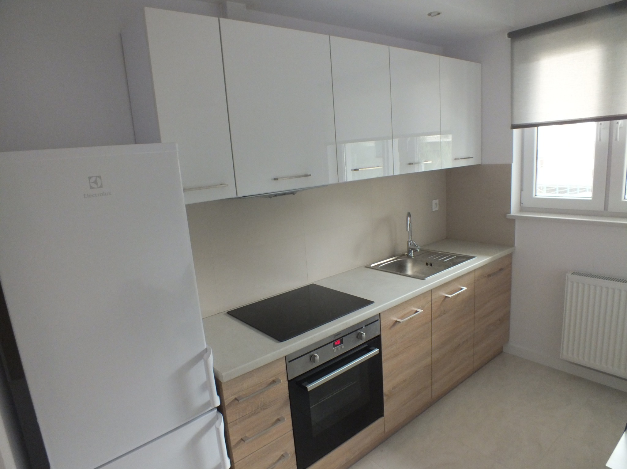 Jasna kuchnia z białymi szafkami górnymi i drewnopodobnymi dolnymi. Widoczny sprzęt AGD: lodówka Electrolux, płyta indukcyjna i piekarnik. Okno z roletą w tle.