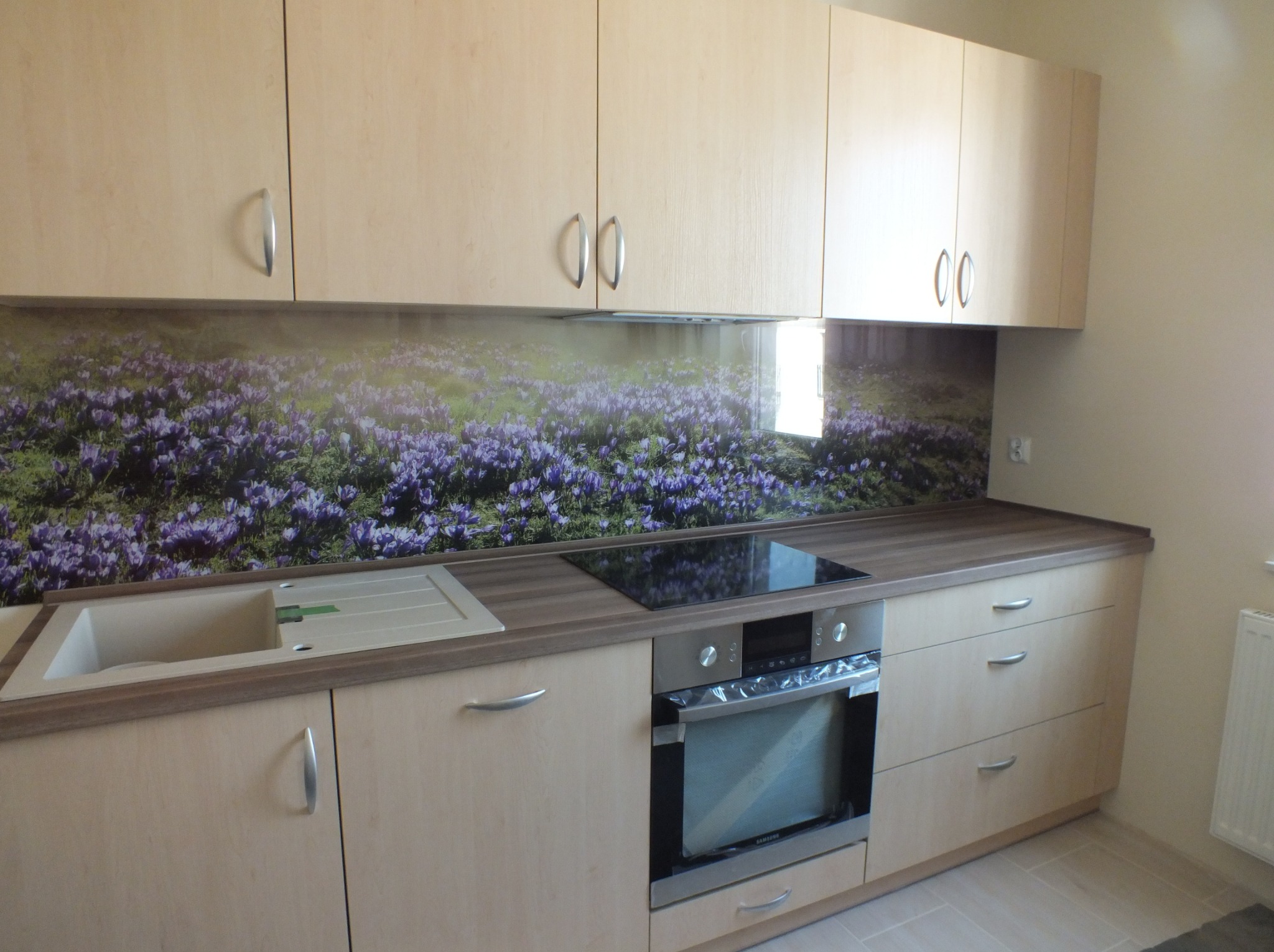 Jasna zabudowa kuchenna z frontami z drewna, srebrne uchwyty, szklany panel nad blatem z motywem fioletowych krokusów, płyta indukcyjna i piekarnik.