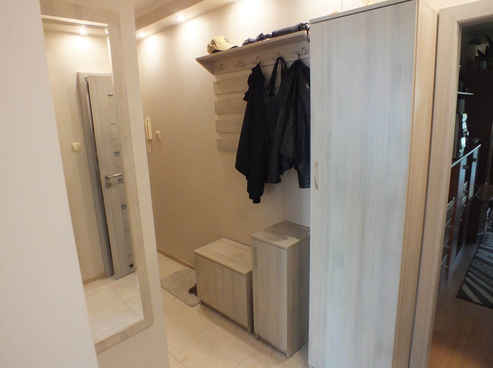 Jasna szafa z wieszakami i półką na buty w przedpokoju, widok z perspektywy lustra. Ubrania wiszą na wieszakach, a na półce leży czapka.