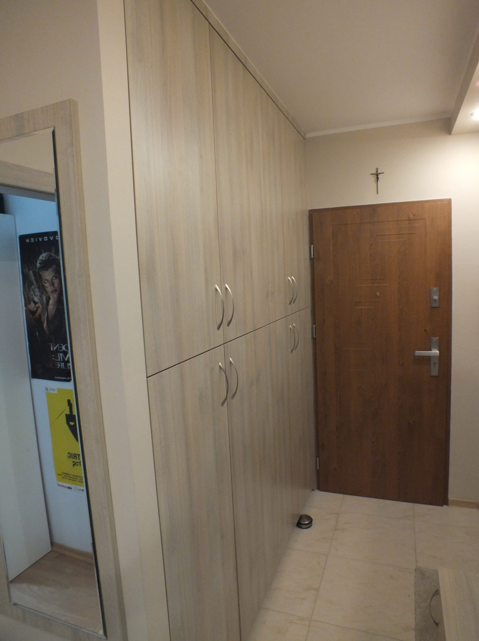 Jasna szafa wnękowa z czterema rzędami uchwytów, widoczna obok lustra i brązowych drzwi wejściowych z krzyżem na ścianie. Neutralna kolorystyka wnętrza.