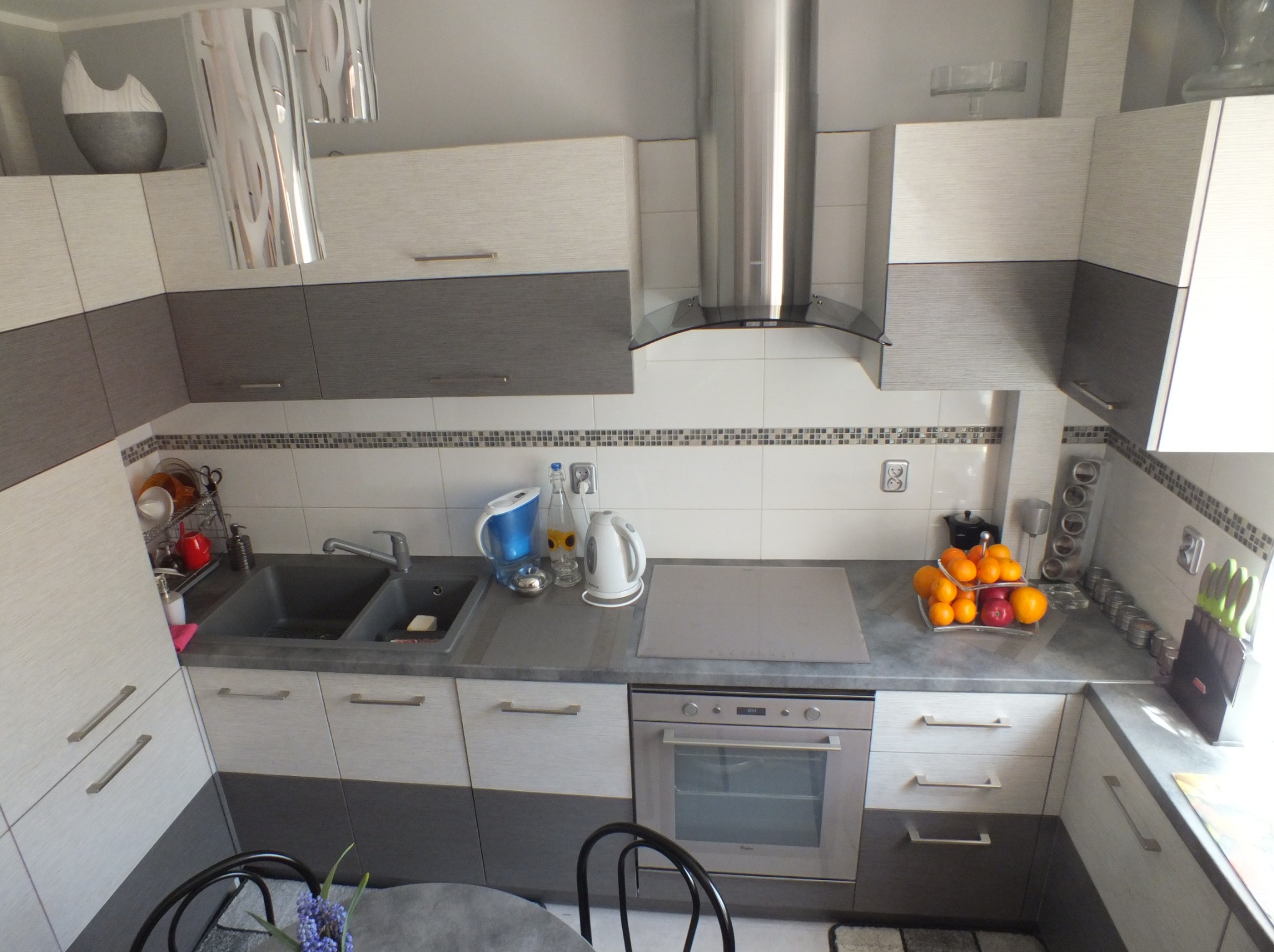 Dwukolorowa zabudowa kuchenna z szarymi dolnymi szafkami i białymi górnymi, uzupełniona o srebrny okap i dekoracyjne akcesoria. Widok na blat z owocami i kwiatami.