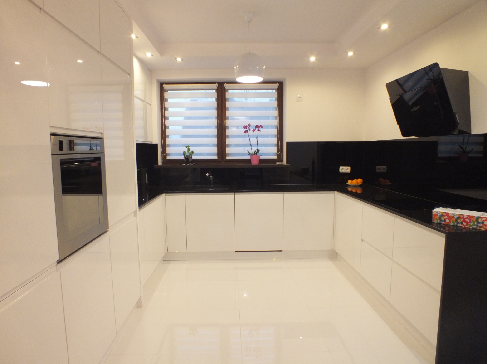 Biała, nowoczesna kuchnia na wymiar z czarnym blatem i oknem z żaluzjami dzień-noc. Minimalistyczny design z połyskującymi frontami i oświetleniem punktowym.