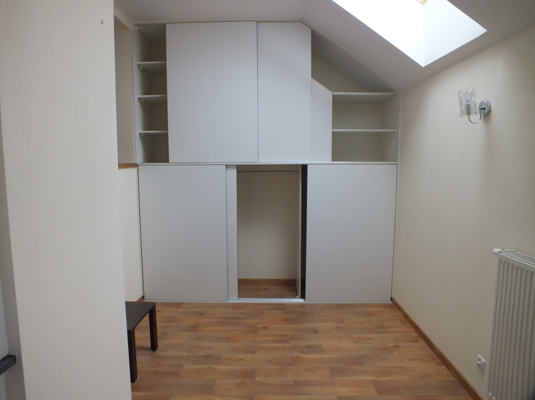 Biała szafa wnękowa z przesuwnymi drzwiami, dopasowana do skosu dachu na poddaszu. Widoczne półki i drewniana podłoga. Oświetlenie górne.