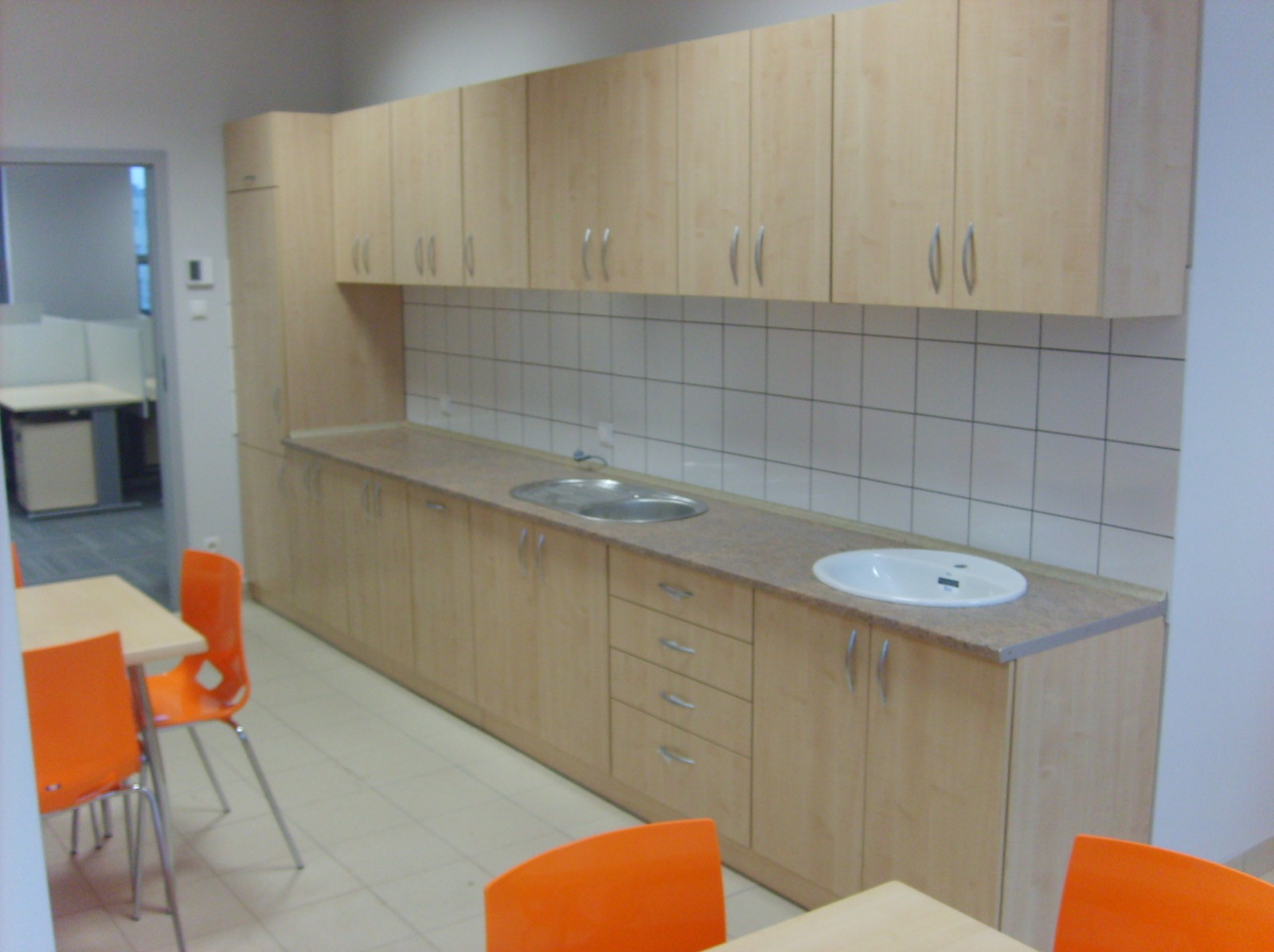 Jasne, drewniane szafki kuchenne z dwoma zlewozmywakami i białymi płytkami w tle. Widoczne pomarańczowe krzesła i stół. Ujęcie z perspektywy lekko z dołu.