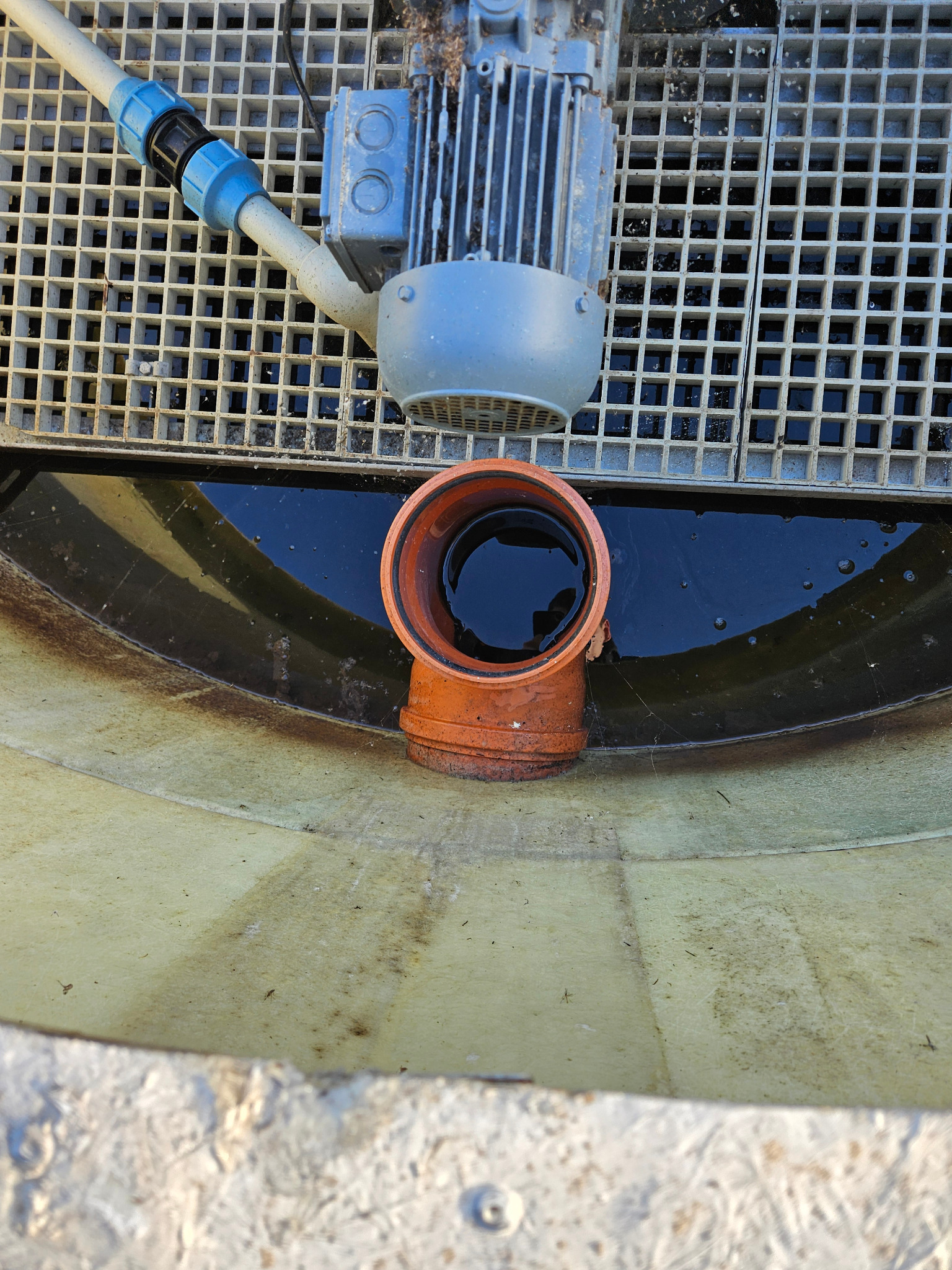 Fragment instalacji oczyszczalni ścieków z pomarańczową rurą i silnikiem, widoczne elementy kraty i zbiornika. Ujęcie z góry, oświetlenie naturalne.