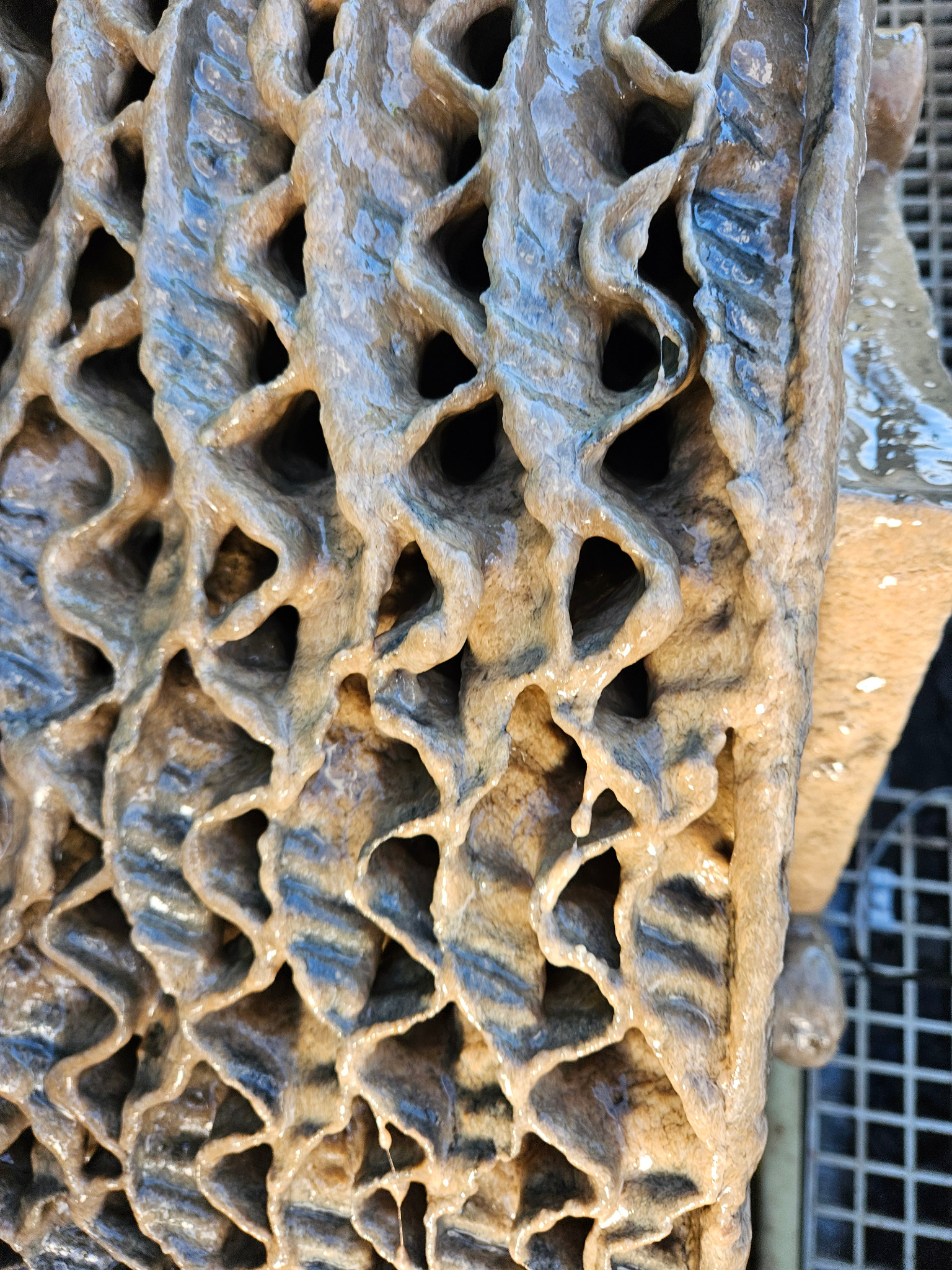 Żebrowana struktura z otworami, prawdopodobnie element drenażowy oczyszczalni ścieków, pokryta wilgotną, brązową substancją. Widoczna kratka w tle.