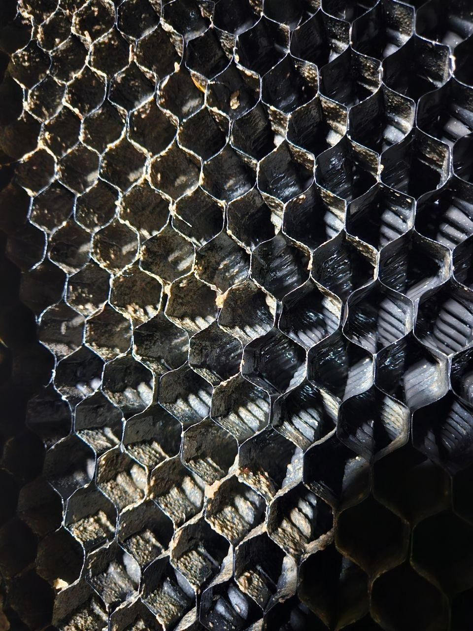 Zbliżenie na strukturę filtra biologicznego oczyszczalni ścieków. Widoczne heksagonalne komory wypełnione osadem, oświetlone z góry, tworzące wzór cieni i refleksów.