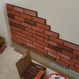 SEB-BUD Sebastian Żebczyński - Częściowo ułożona dekoracyjna ściana z cegły klinkierowej w pomieszczeniu. Widoczne narzędzia i materiały budowlane: wiadro z zaprawą, pędzle, opakowanie z cegłami.