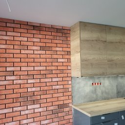 SEB-BUD Sebastian Żebczyński - Ceglana ściana w kuchni z drewnianymi szafkami i szarą płytą. Widoczne gniazdka elektryczne i fragment blatu. Detal wykończenia wnętrza.