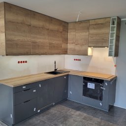 SEB-BUD Sebastian Żebczyński - Nowoczesna kuchnia w trakcie montażu: szare szafki dolne z drewnianym blatem, górne szafki z drewnopodobnym wzorem, czarny zlew, płytki podłogowe. Widoczne gniazdka elektryczne.