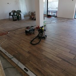 SEB-BUD Sebastian Żebczyński - Układanie drewnopodobnych płytek podłogowych w Olkuszu. Widoczny poziomica, odkurzacz i narzędzia. Duże okno w tle. Prace wykończeniowe w nowym domu.