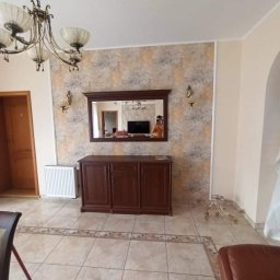 SEB-BUD Sebastian Żebczyński - Elegancki salon z drewnianą komodą pod lustrem w ozdobnej ramie, wzorzysta tapeta, kremowe płytki podłogowe z dekoracyjnym obramowaniem i żyrandol w stylu retro.