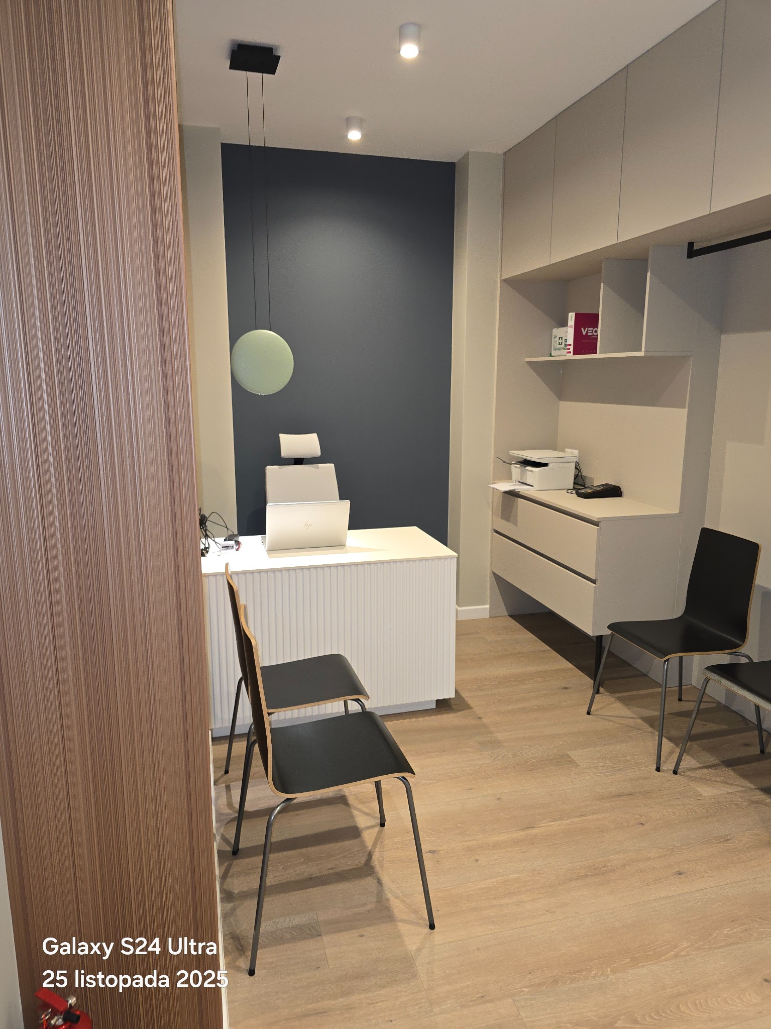 Nowoczesne biuro po remoncie: minimalistyczne meble, drewniana podłoga, lampa wisząca. Stonowana kolorystyka ścian i zabudowy meblowej tworzy elegancką przestrzeń.