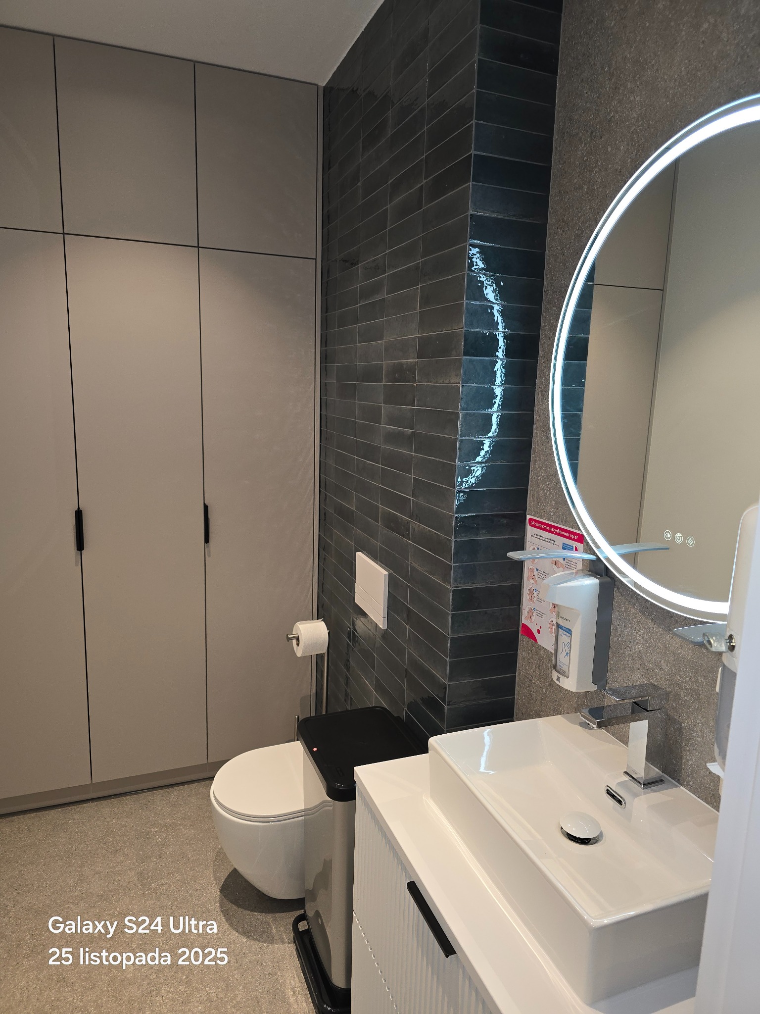 Nowoczesna łazienka z ciemnymi, błyszczącymi płytkami ułożonymi w cegiełkę, okrągłym lustrem z podświetleniem LED i minimalistyczną białą umywalką z szafką.