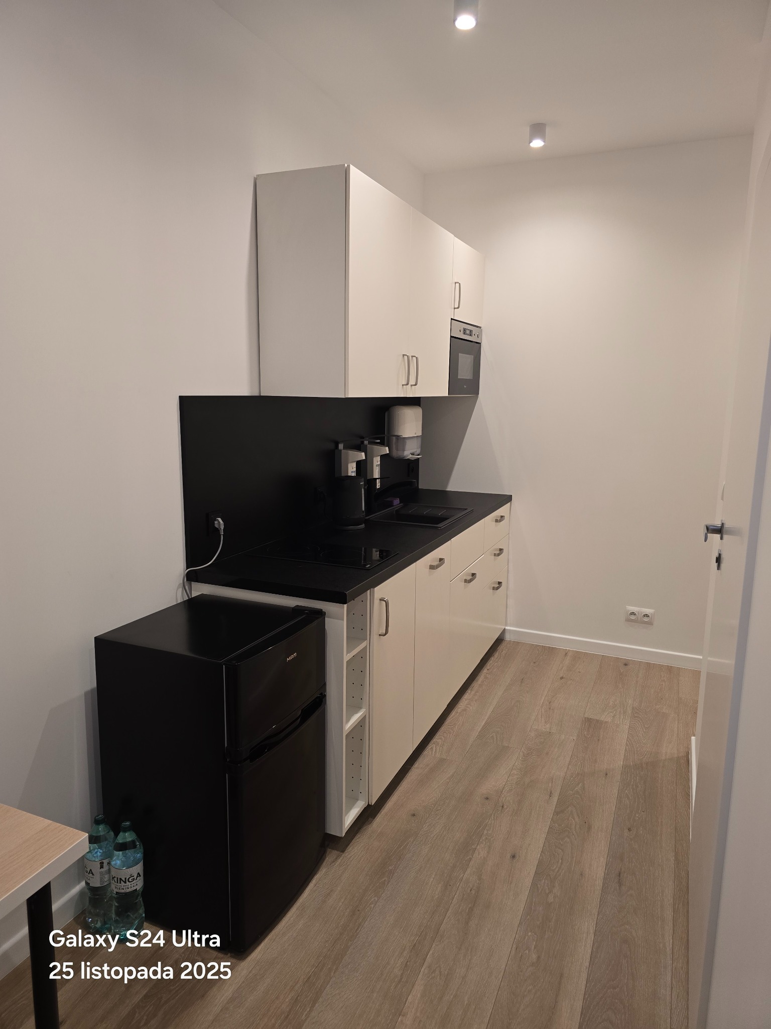 Nowoczesna, minimalistyczna kuchnia z białymi szafkami, czarnym blatem i małą czarną lodówką. Jasne drewniane panele podłogowe. Widok na fragment otwartego pomieszczenia.