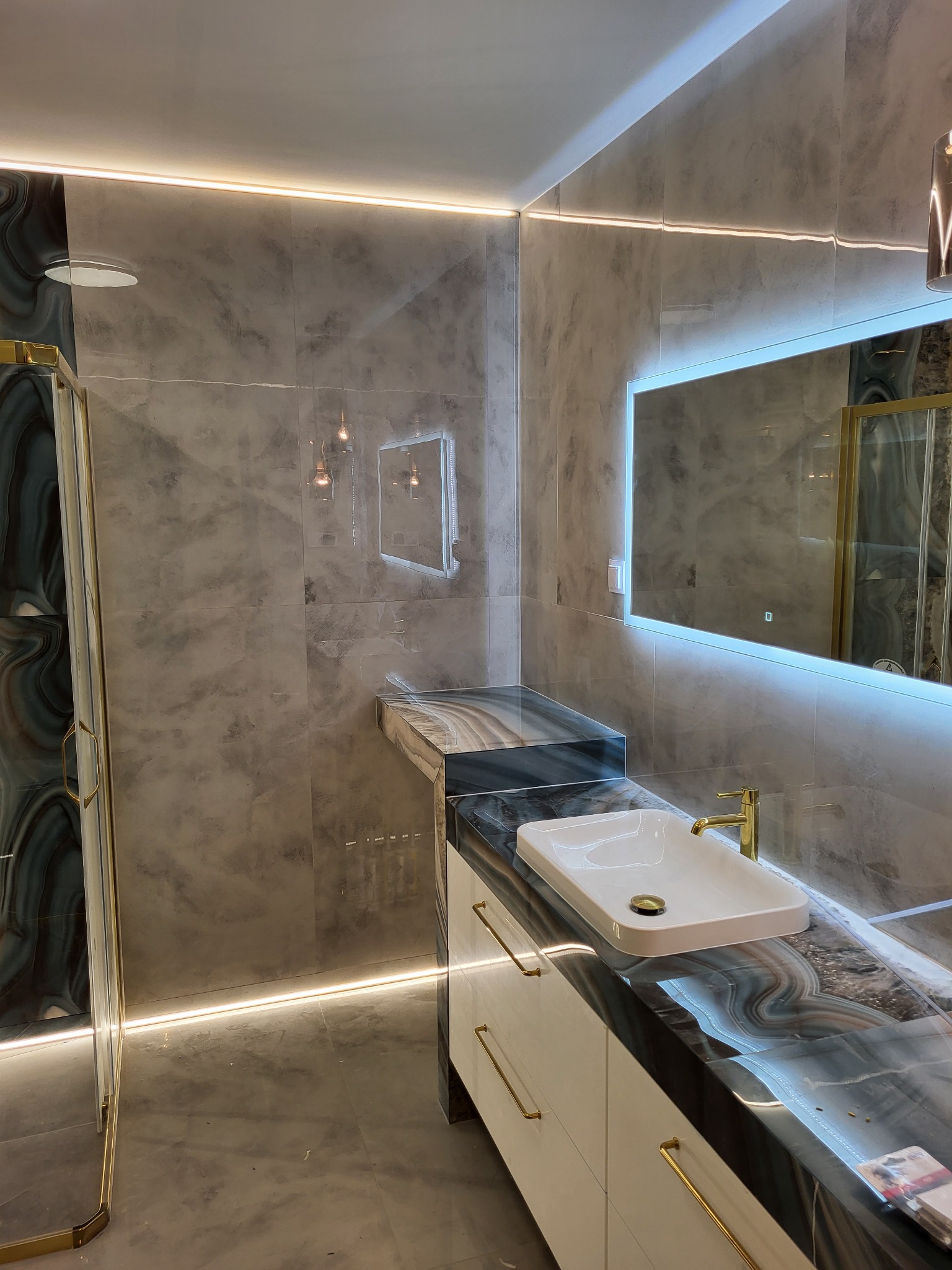 Elegancka łazienka z marmurowymi płytkami, podświetlanym lustrem i złotymi akcentami. Nowoczesna umywalka nablatowa i oświetlenie LED tworzą luksusowy klimat.