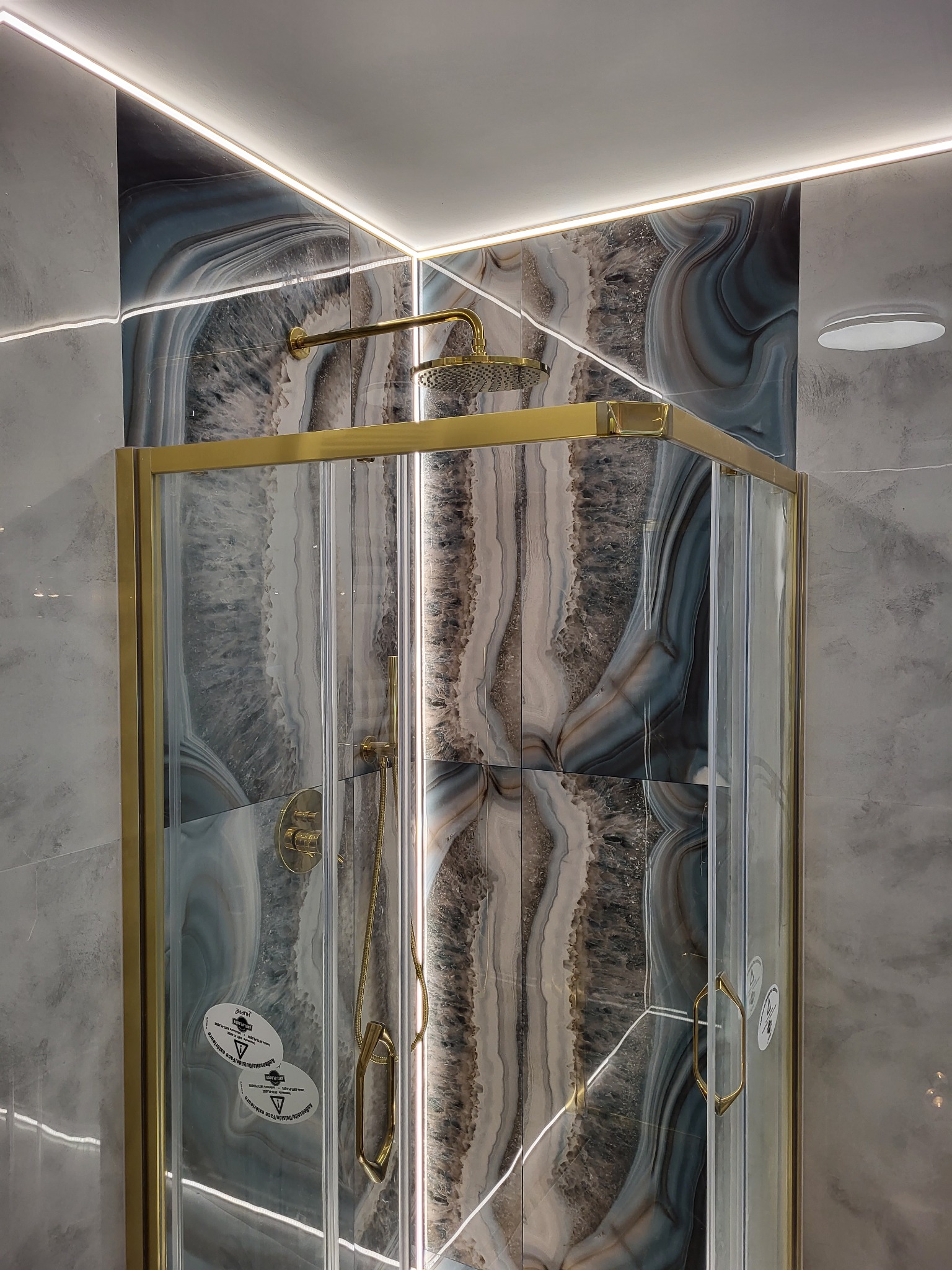 Kabina prysznicowa z złotymi detalami i panelami imitującymi agat, oświetlona listwą LED, na tle szarej ściany. Widoczne naklejki informacyjne na szkle.