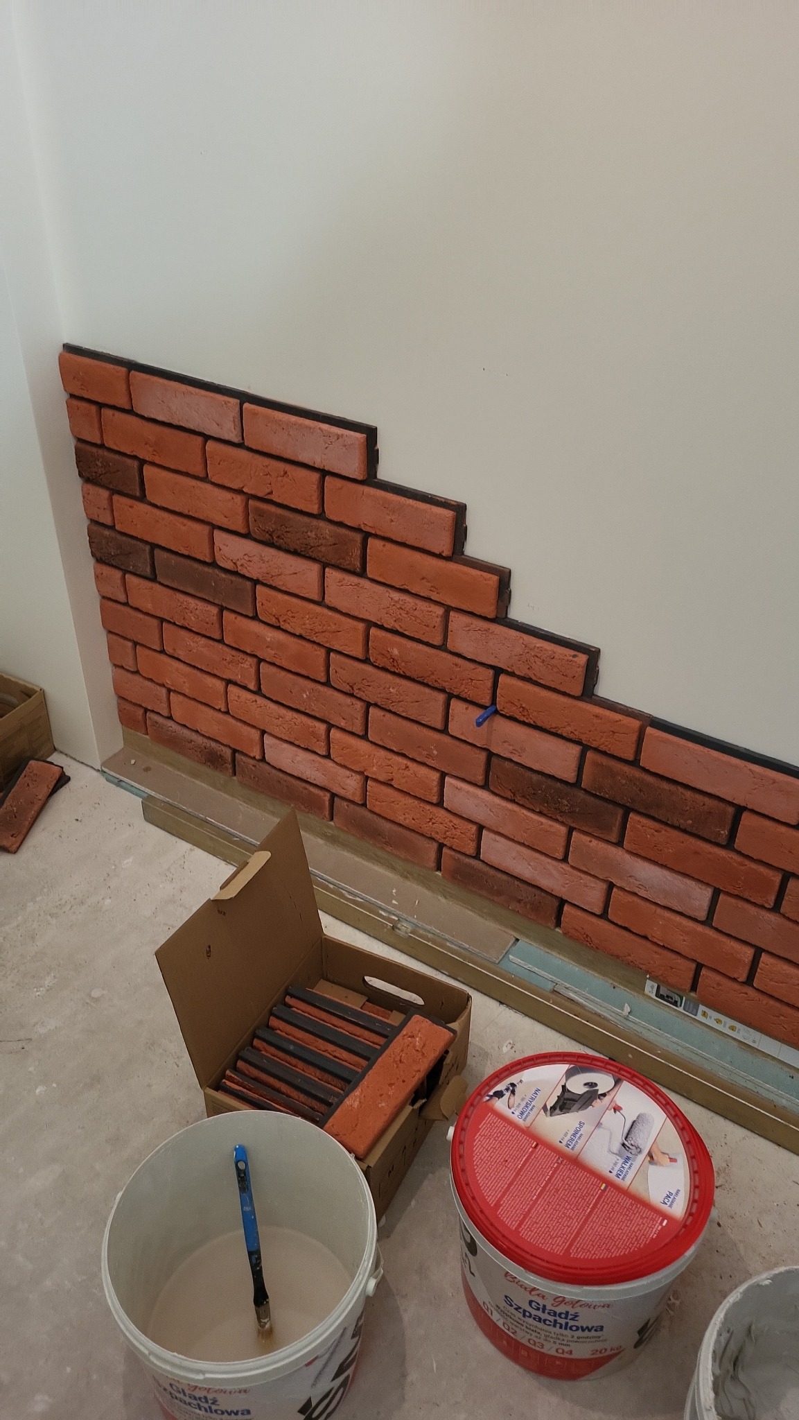 Częściowo ułożona dekoracyjna ściana z cegły klinkierowej w pomieszczeniu. Widoczne narzędzia i materiały budowlane: wiadro z zaprawą, pędzle, opakowanie z cegłami.