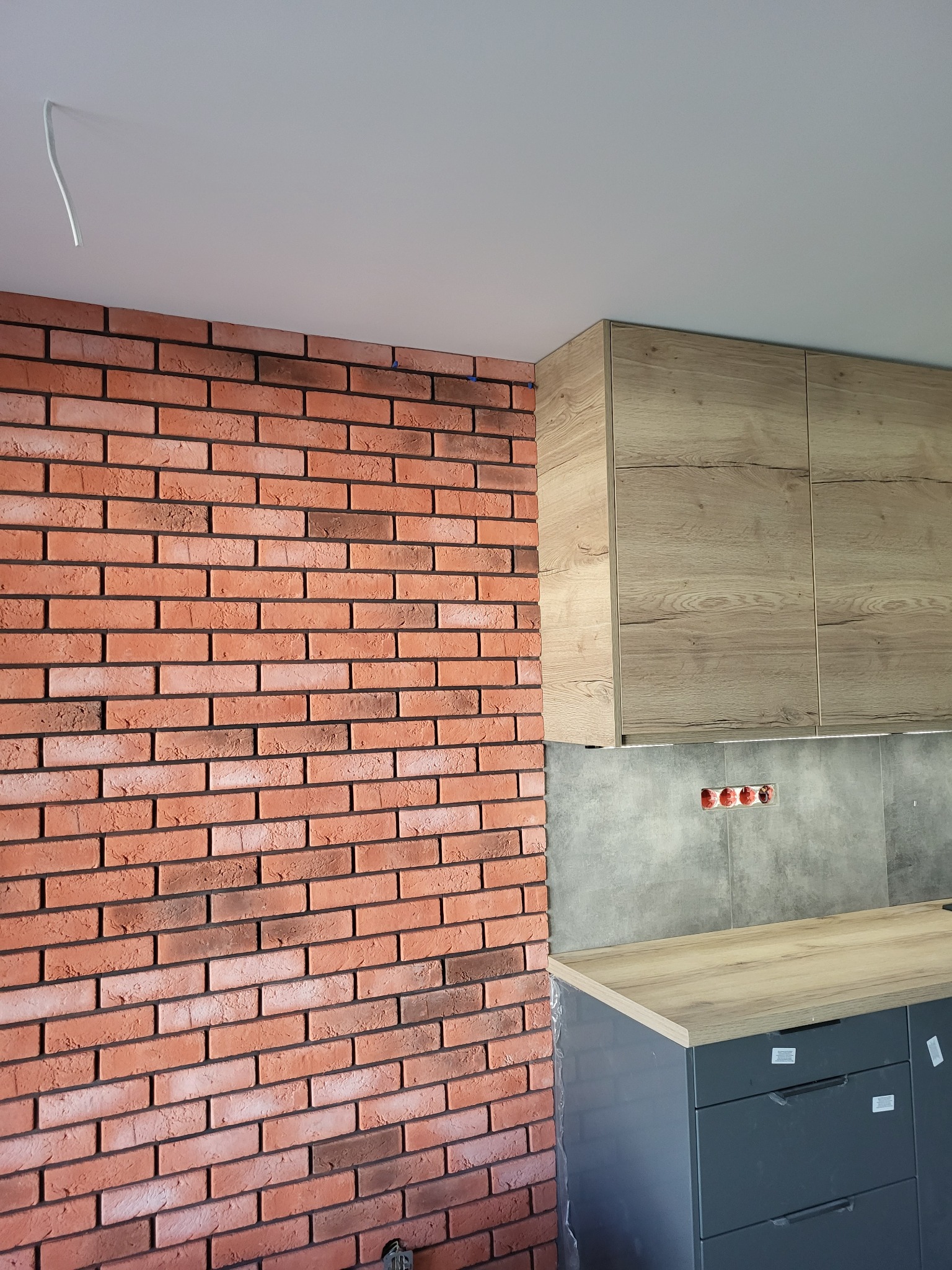 Ceglana ściana w kuchni z drewnianymi szafkami i szarą płytą. Widoczne gniazdka elektryczne i fragment blatu. Detal wykończenia wnętrza.