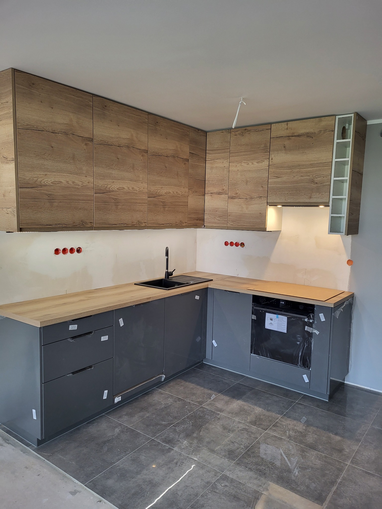 Nowoczesna kuchnia w trakcie montażu: szare szafki dolne z drewnianym blatem, górne szafki z drewnopodobnym wzorem, czarny zlew, płytki podłogowe. Widoczne gniazdka elektryczne.
