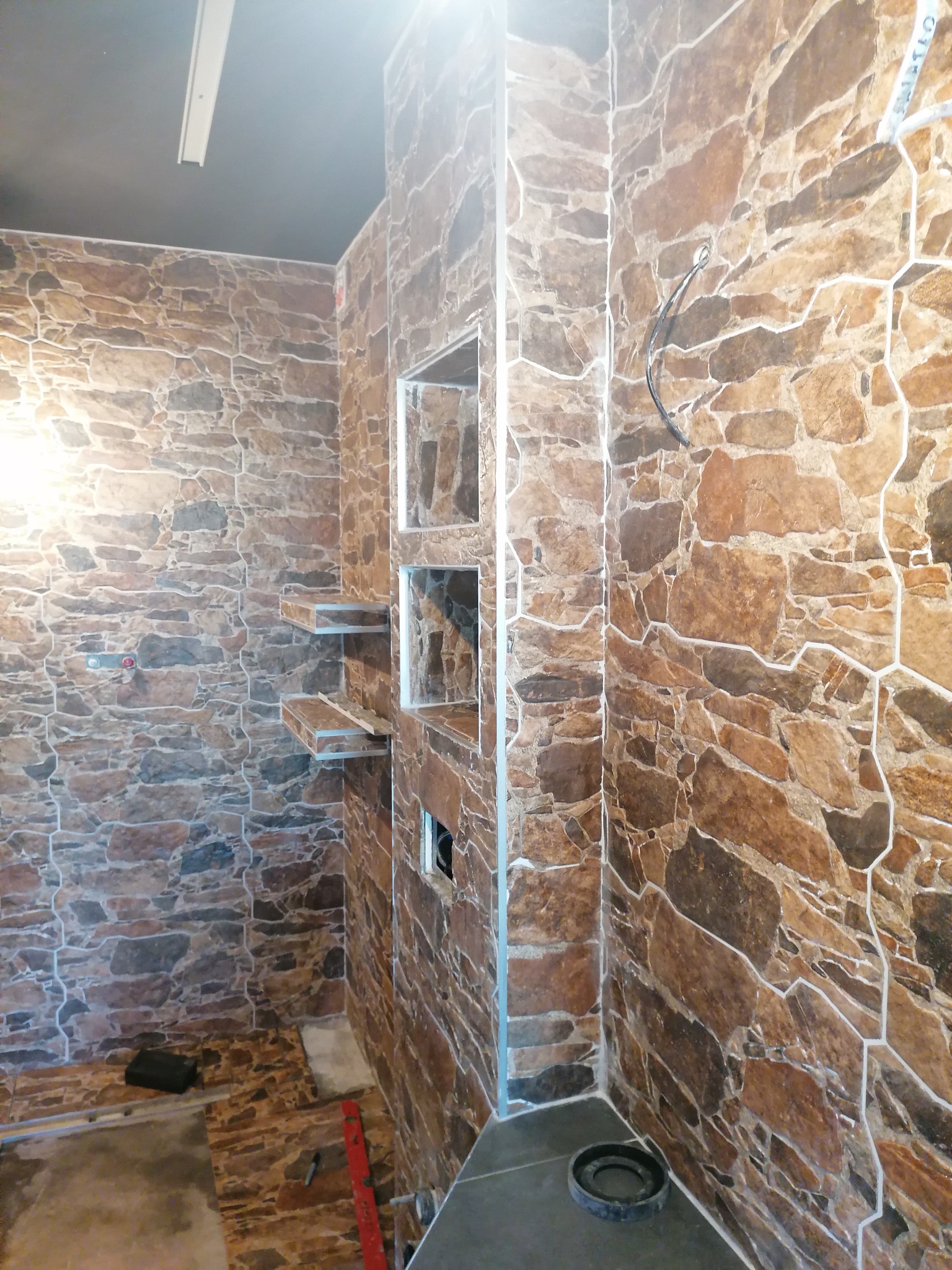 Łazienka w trakcie wykończenia: ściany i podłoga wyłożone płytkami imitującymi kamień, widoczne półki wnękowe i wystające przewody elektryczne.