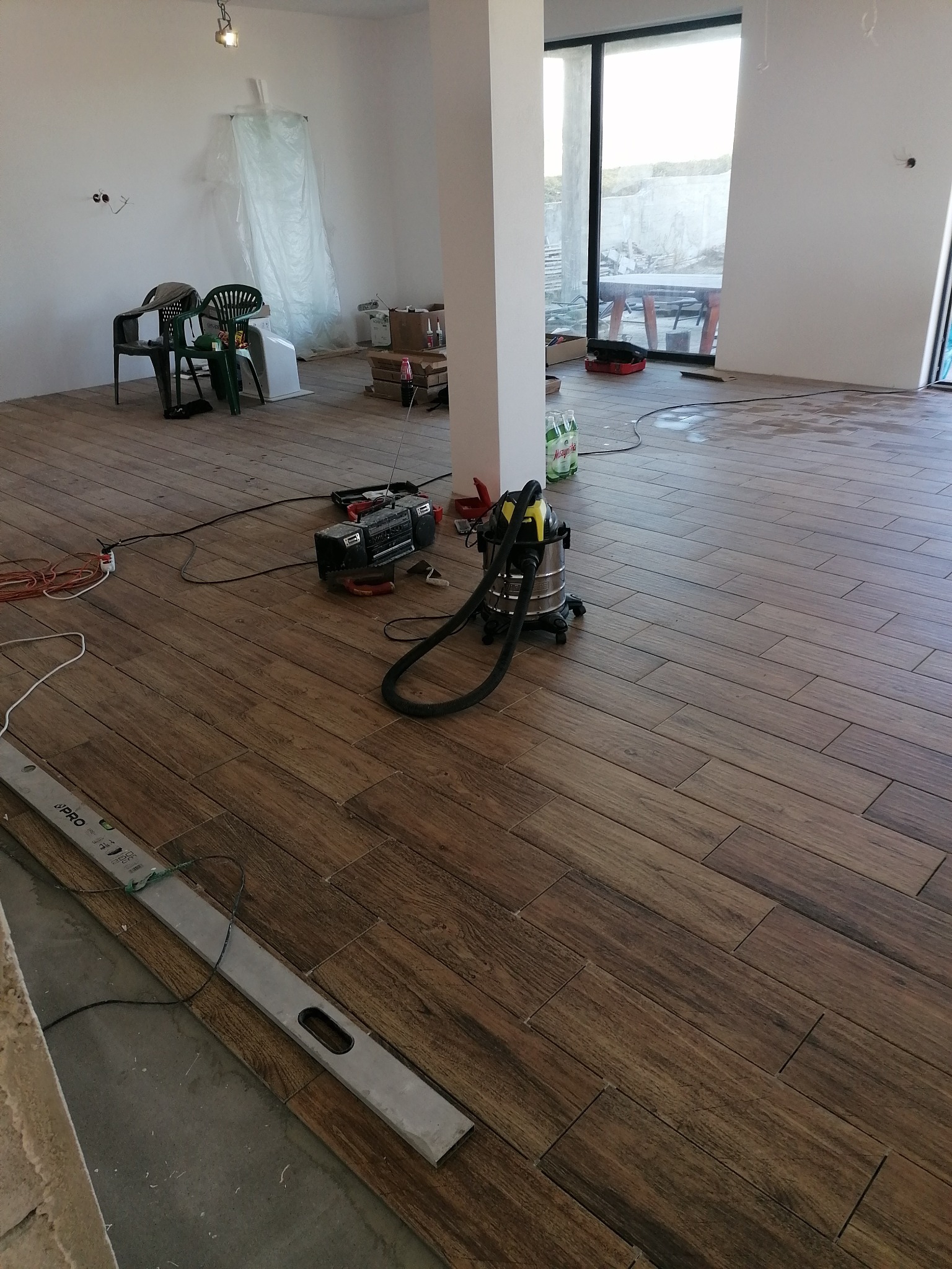 Układanie drewnopodobnych płytek podłogowych w Olkuszu. Widoczny poziomica, odkurzacz i narzędzia. Duże okno w tle. Prace wykończeniowe w nowym domu.