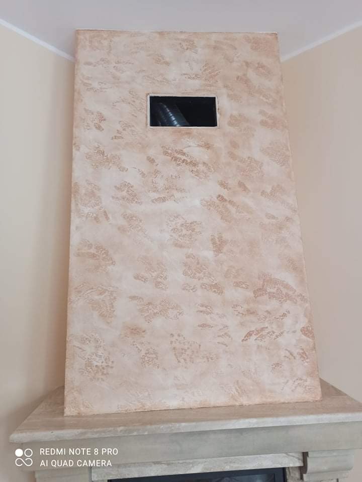 Otynkowana obudowa kominka z otworem wentylacyjnym, dekoracyjna faktura w odcieniach beżu i brązu, widok z dołu, fragment marmurowego blatu kominka.