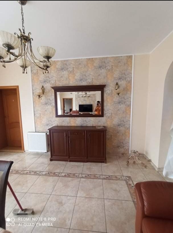 Elegancki salon z drewnianą komodą pod lustrem w ozdobnej ramie, wzorzysta tapeta, kremowe płytki podłogowe z dekoracyjnym obramowaniem i żyrandol w stylu retro.