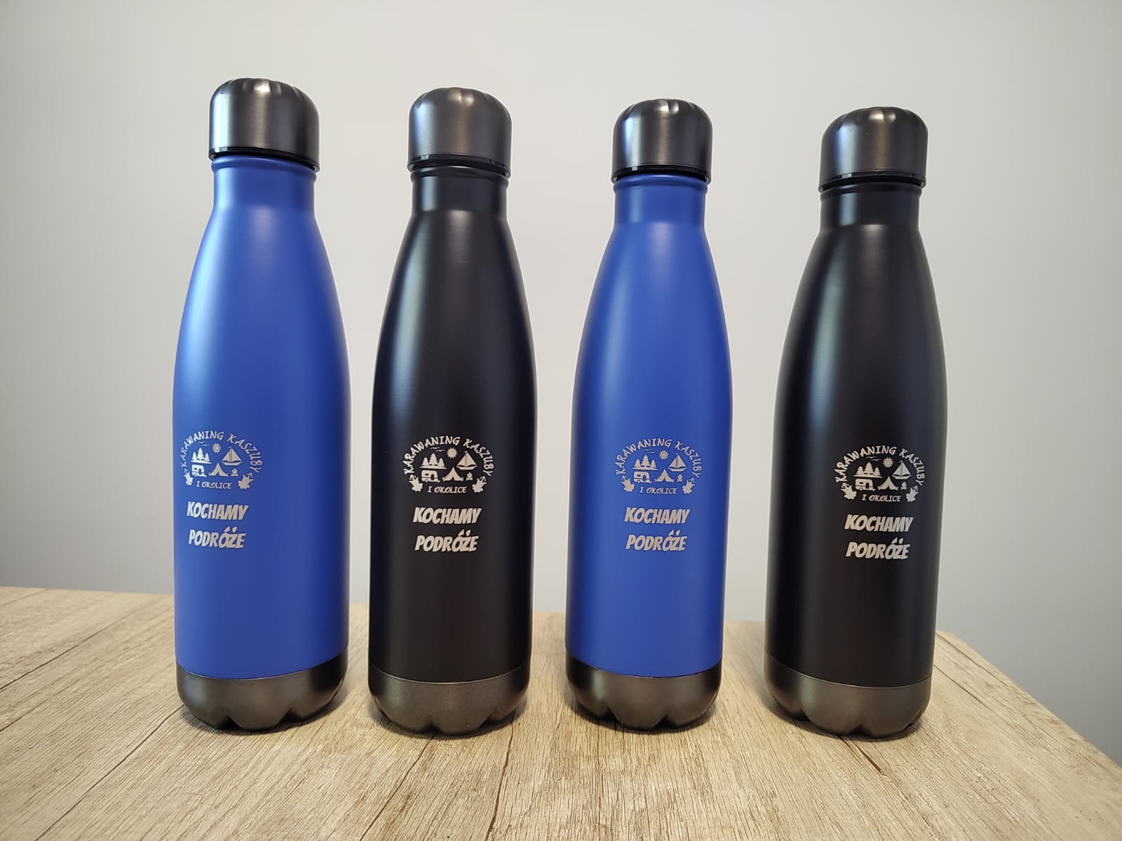 Cztery metalowe butelki (dwie niebieskie i dwie czarne) z nadrukiem 'Karawaning Kaszub i okolice. Kochamy podróże' stoją na drewnianym blacie.