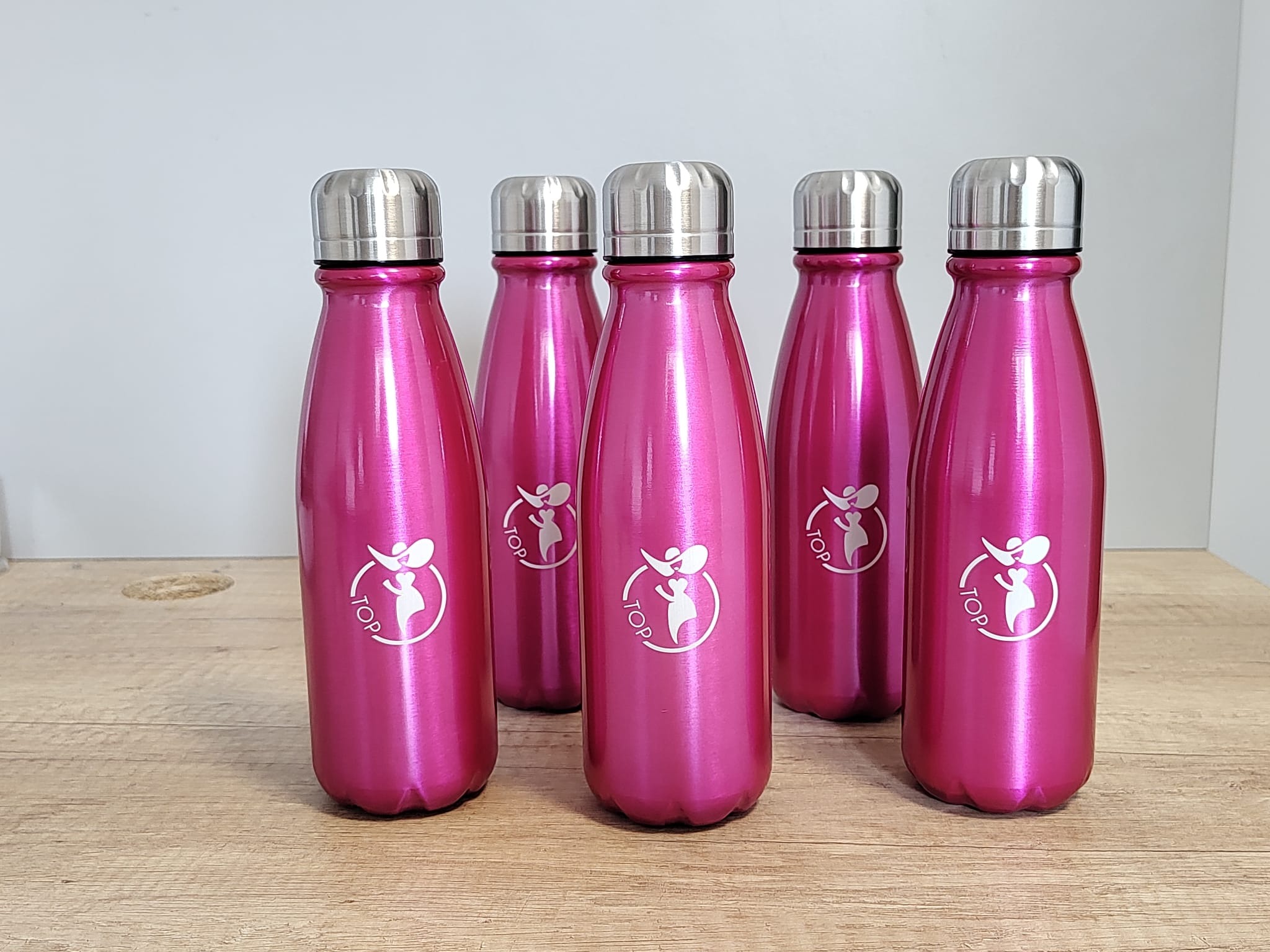 Rząd pięciu metalowych, różowych butelek z logo firmy na drewnianym blacie, na tle szarej ściany. Butelki mają srebrne nakrętki.