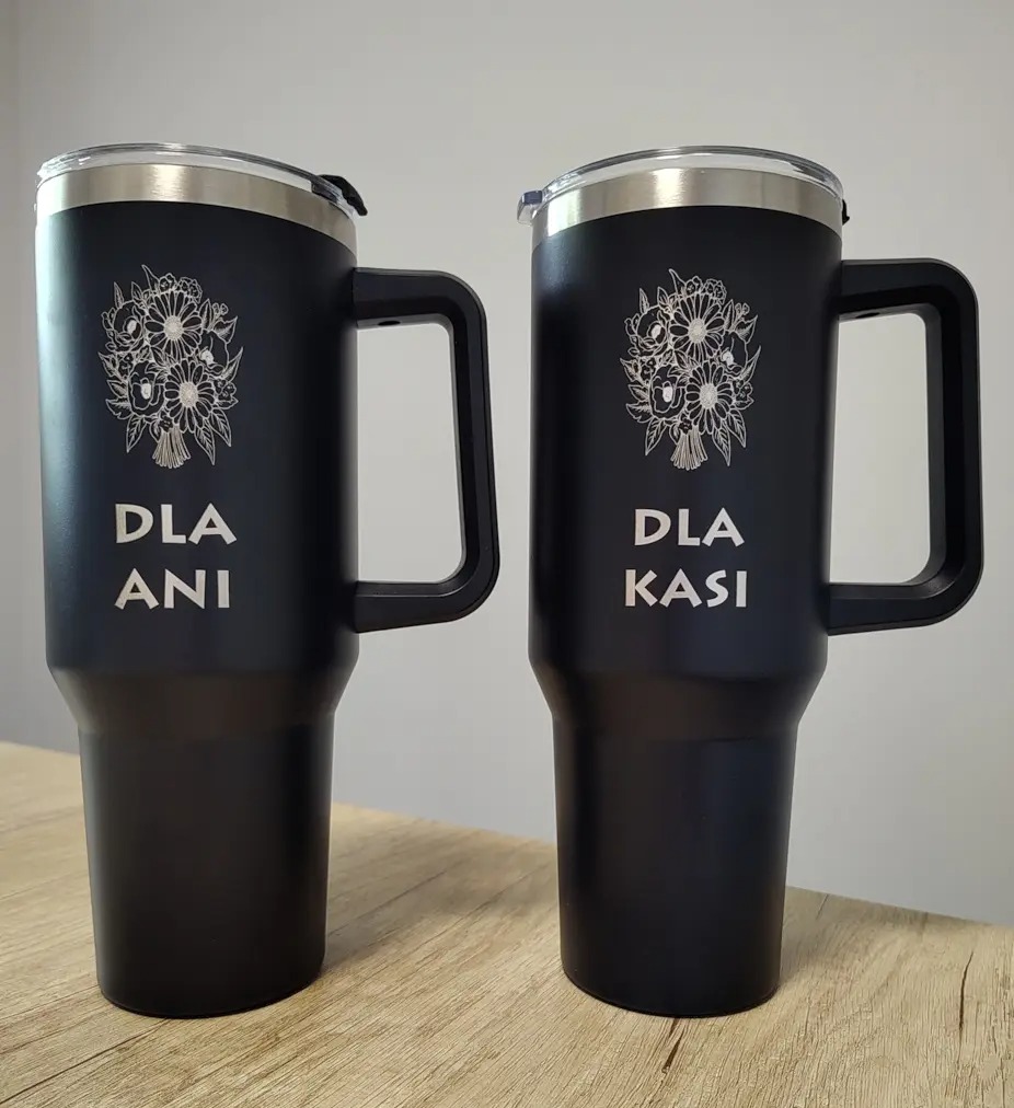 Dwa czarne kubki termiczne z grawerem kwiatów i imion 'Dla Ani' i 'Dla Kasi', stojące na drewnianym blacie, prezentujące spersonalizowane upominki.