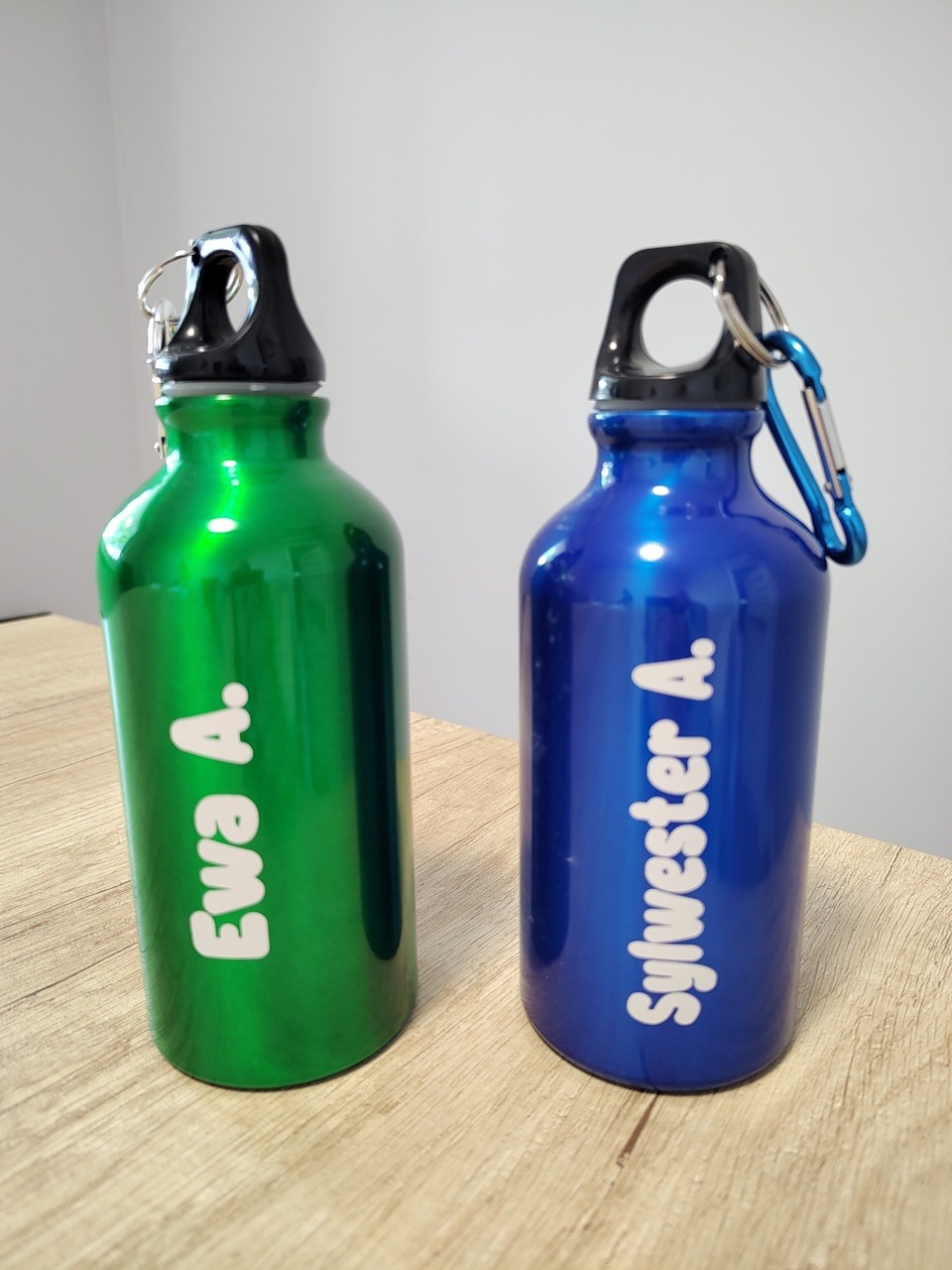 Dwie metalowe butelki na wodę: zielona z napisem 'Ewa A.' i niebieska z napisem 'Sylwester A.', stojące na drewnianym blacie, z karabińczykami przy nakrętkach.