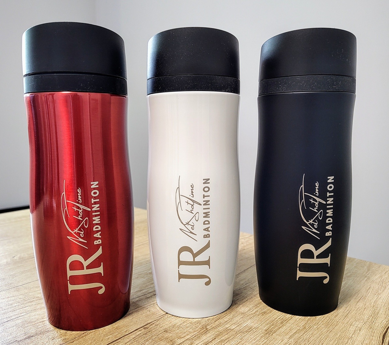 Trzy metalowe kubki termiczne z logo JR Badminton w kolorach: czerwonym, białym i czarnym, stojące na drewnianym blacie w Kamionkach. Personalizacja gadżetów reklamowych.