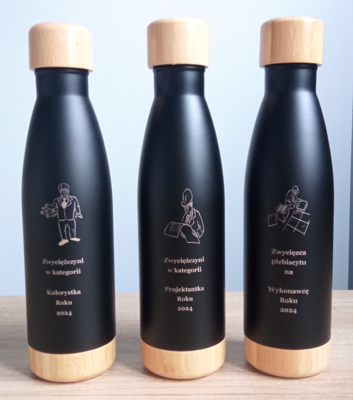 Trzy czarne butelki z bambusowymi elementami, każda z unikalnym grawerem: Kolorystka Roku, Projektantka Roku, Wykonawca Roku 2024. Nagrody plebiscytu.