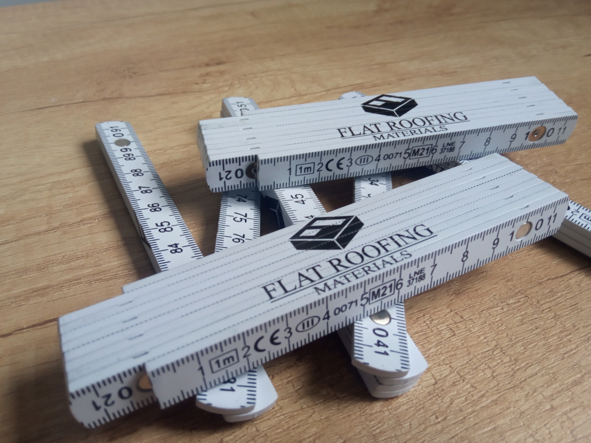Rozłożone, białe miarki składane z nadrukiem 'FLAT ROOFING MATERIALS' na drewnianym tle. Precyzyjne narzędzie pomiarowe z logo firmy.