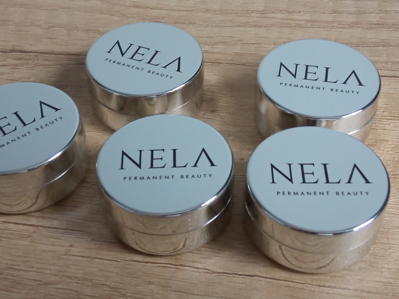 Pięć srebrnych słoiczków z jasnoniebieskimi wieczkami, na których widnieje napis 'NELA PERMANENT BEAUTY' na drewnianym tle. Minimalistyczny design.
