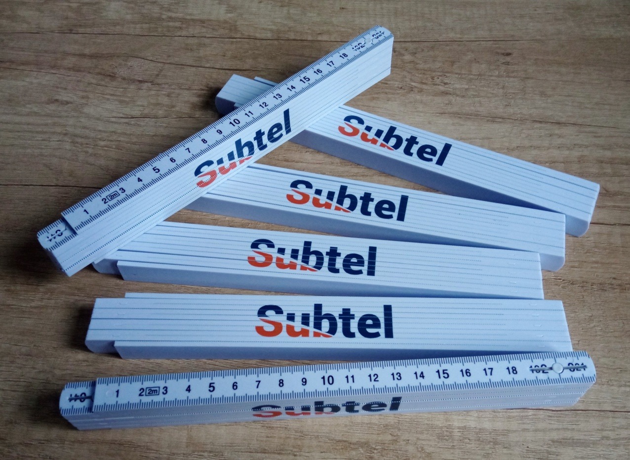 Składana, biała miarka z nadrukowanym logo 'Subtel' w kolorze granatowo-pomarańczowym, ułożona na drewnianym tle. Widoczne podziałki centymetrowe i milimetrowe.