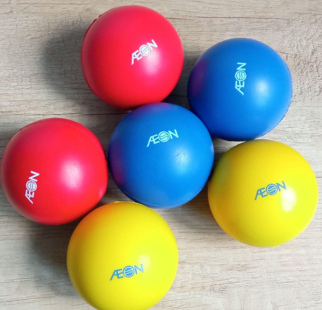 Sześć kolorowych piłek (czerwona, niebieska, żółta) z logo AEON na drewnianym tle. Piłki ułożone blisko siebie, tworząc kompozycję.