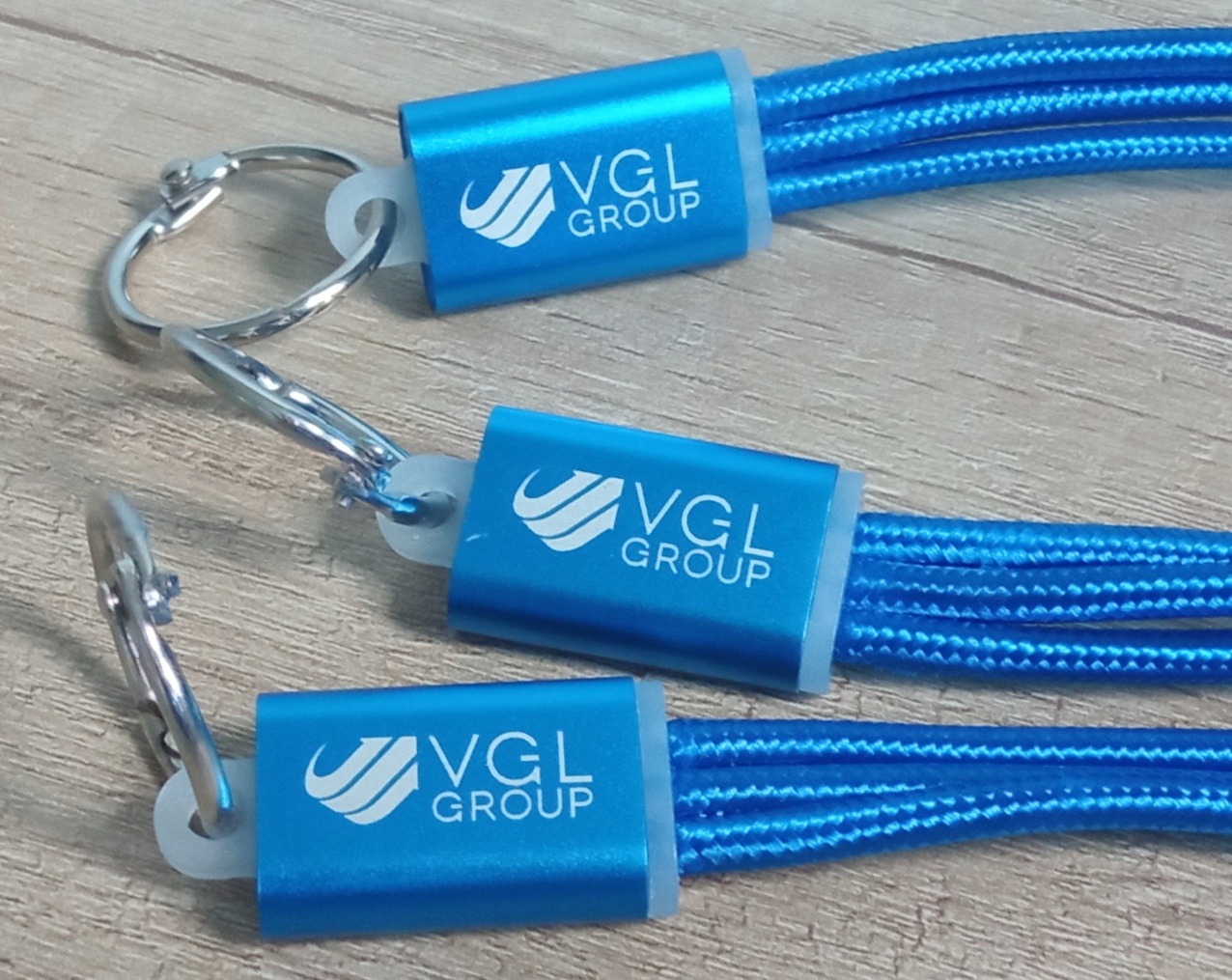 Trzy niebieskie smycze z logo VGL GROUP, ułożone ukośnie na drewnianym tle, z metalowymi karabińczykami. Widoczne detale splotu i metalowych elementów.