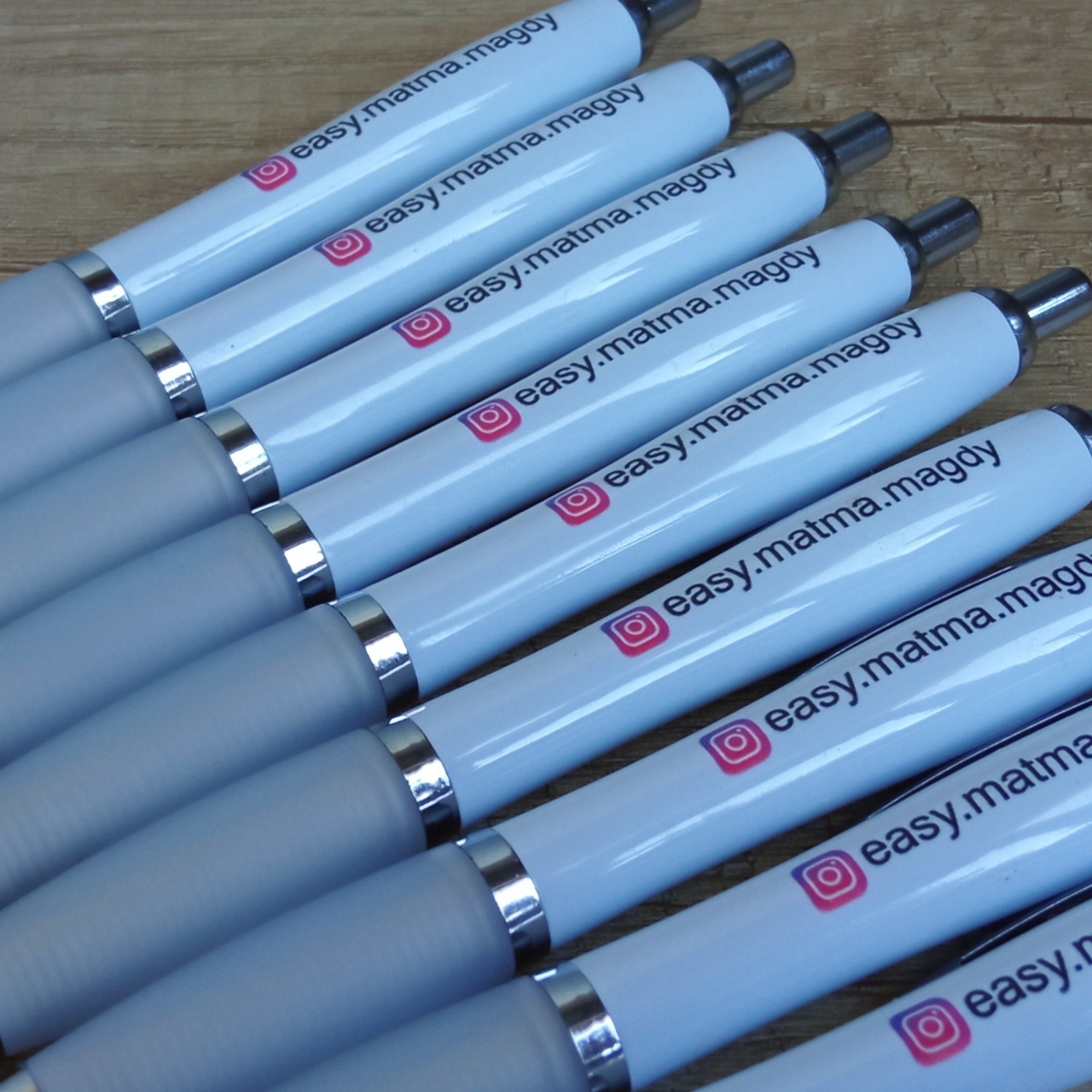 Rząd białych długopisów z szarymi uchwytami, każdy z nadrukowanym logo Instagrama i adresem 'easy.matma.magdy' na drewnianym tle.
