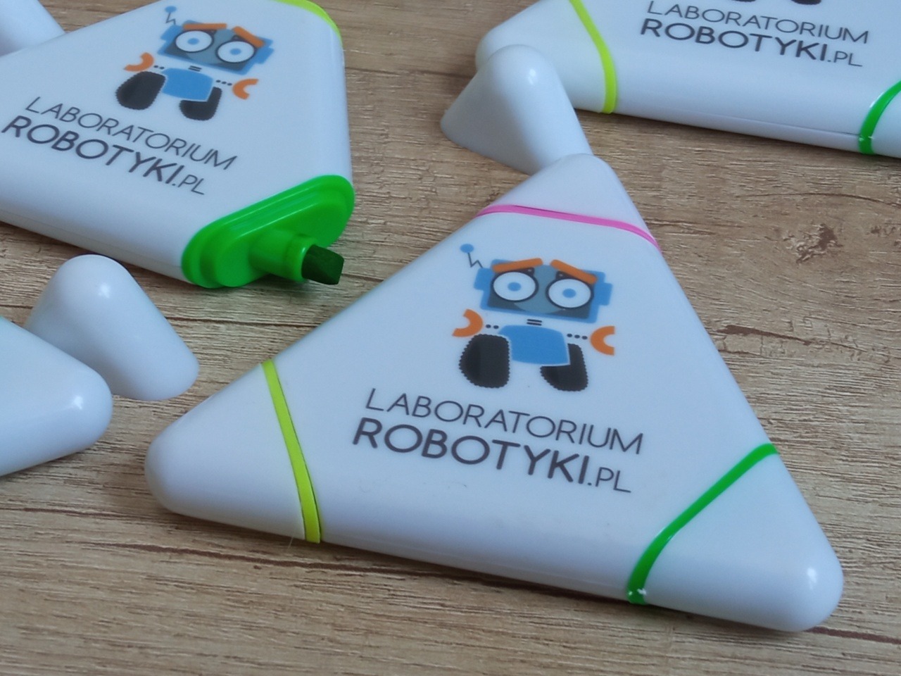 Trójkątne zakreślacze z logo 'Laboratorium Robotyki.pl' i rysunkiem robota na drewnianym tle. Zakreślacze w kolorach: zielony, żółty, różowy.